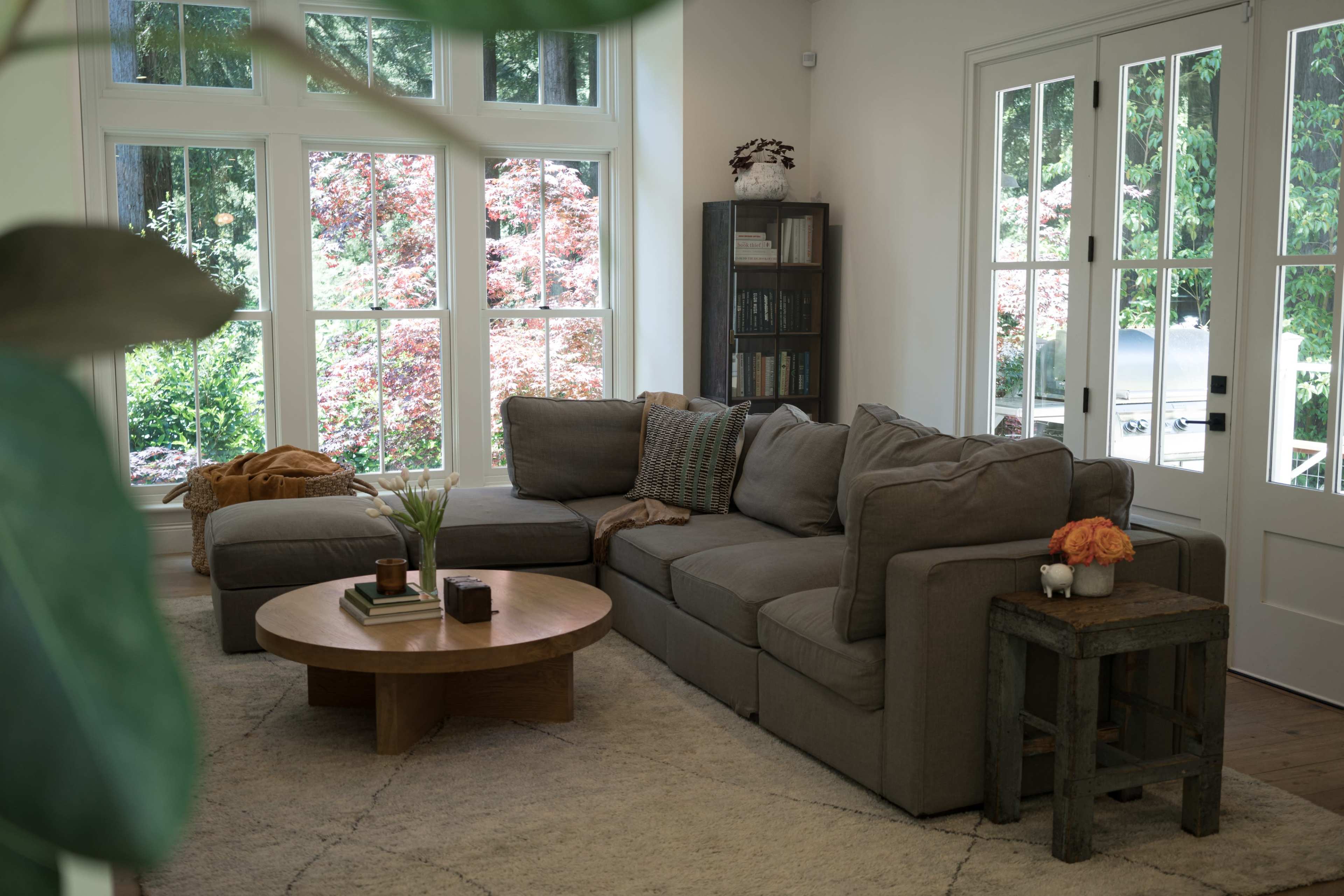 A modern living room features a large sectional sofa, a round wooden coffee table, and large windows showcasing a garden view.