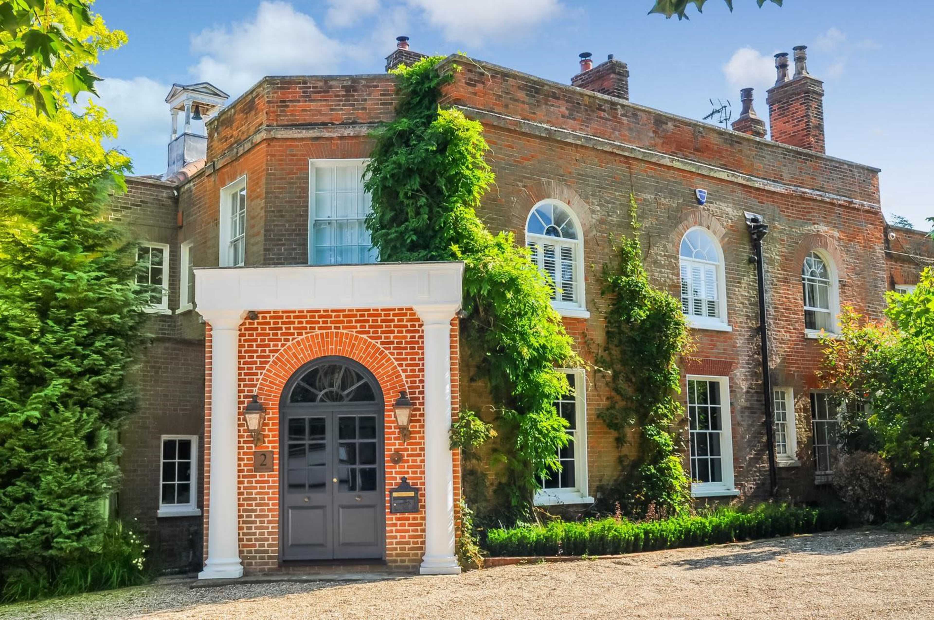 A two-story brick house with large windows and a front entrance framed by decorative columns is surrounded by greenery.