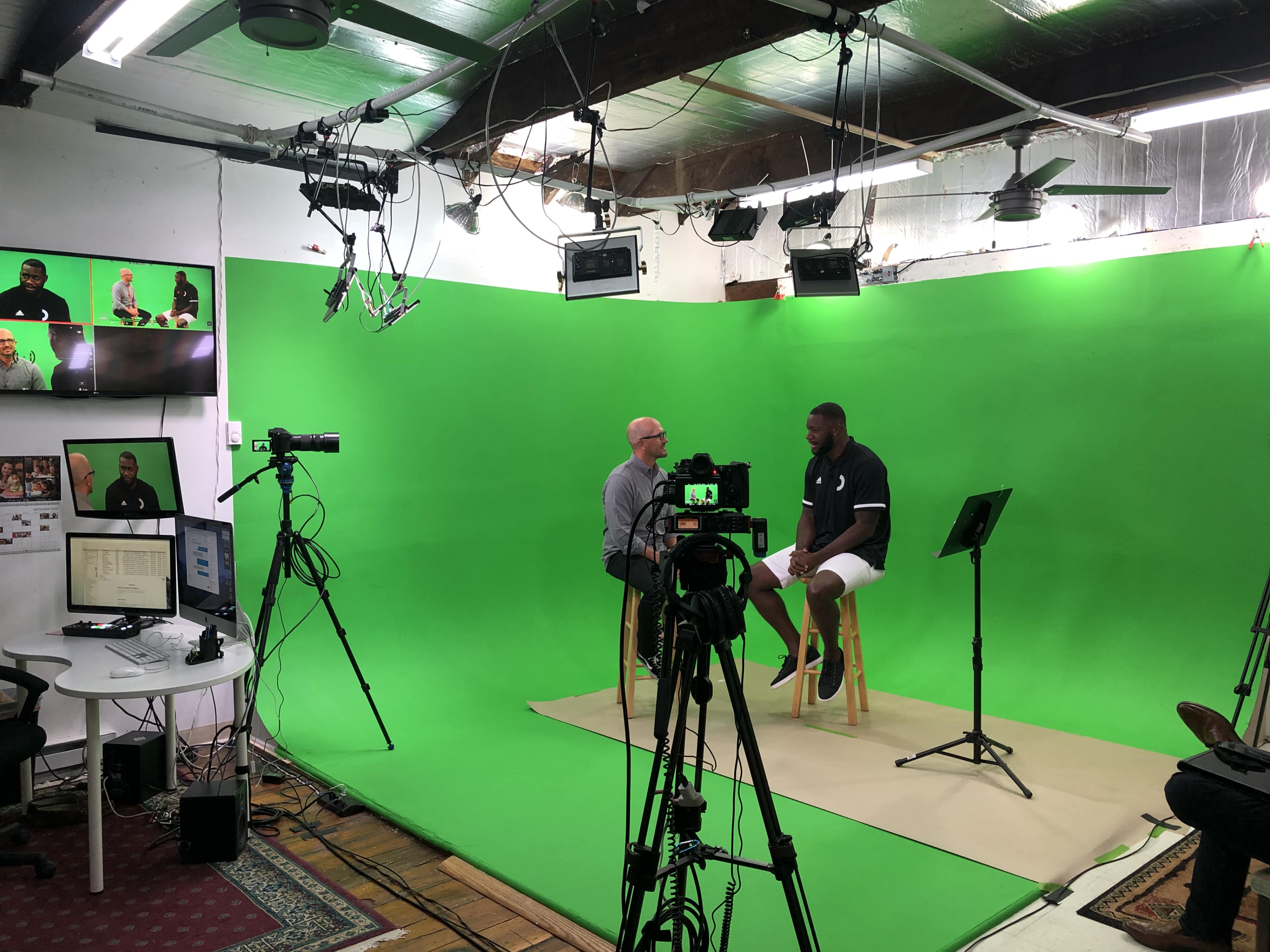 A man sits on a stool in front of a green screen during an interview, while cameras and equipment are arranged around the room.