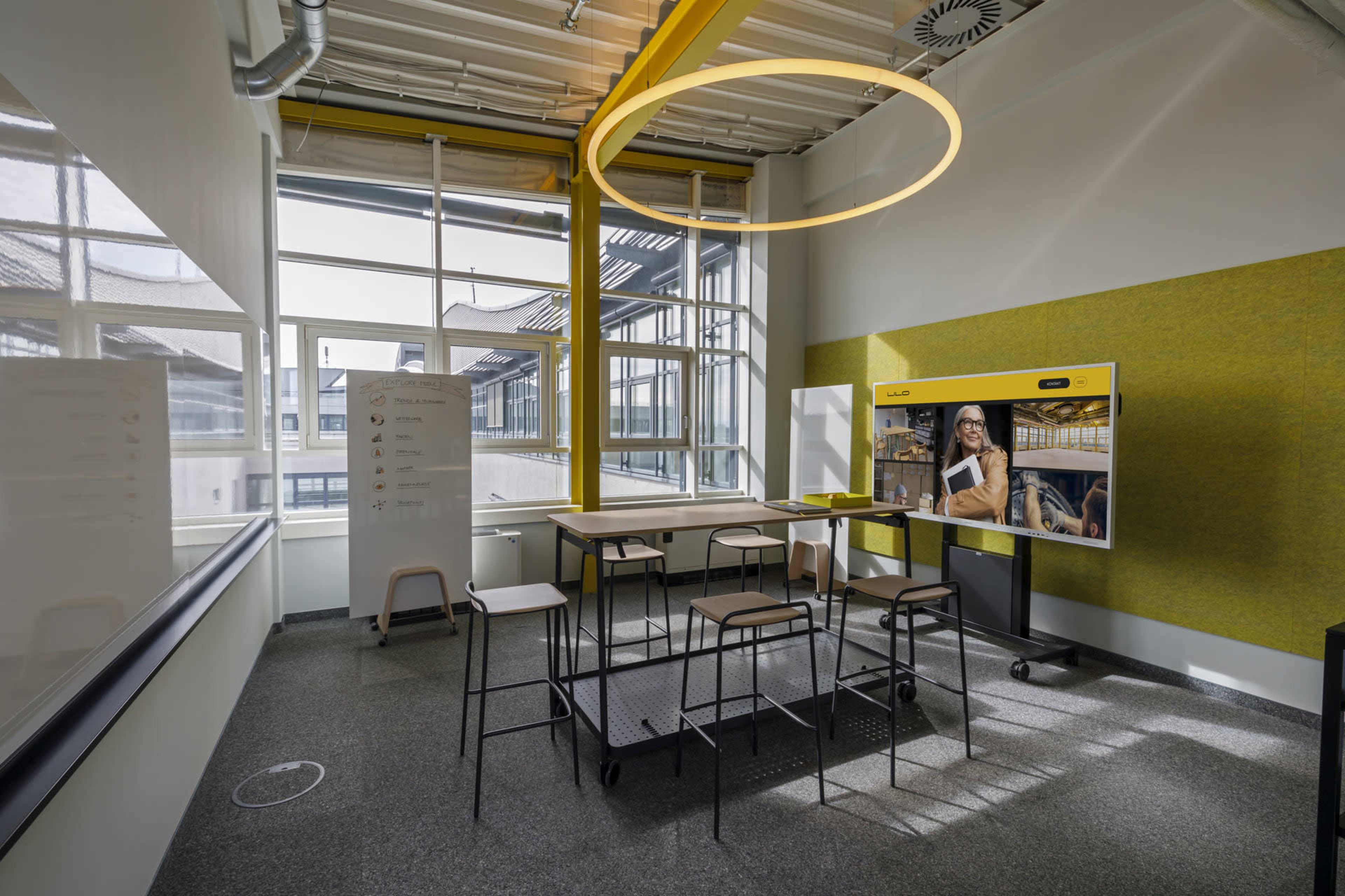 The image shows a modern meeting room with large windows, a wooden table surrounded by stools, and a digital display on one wall.