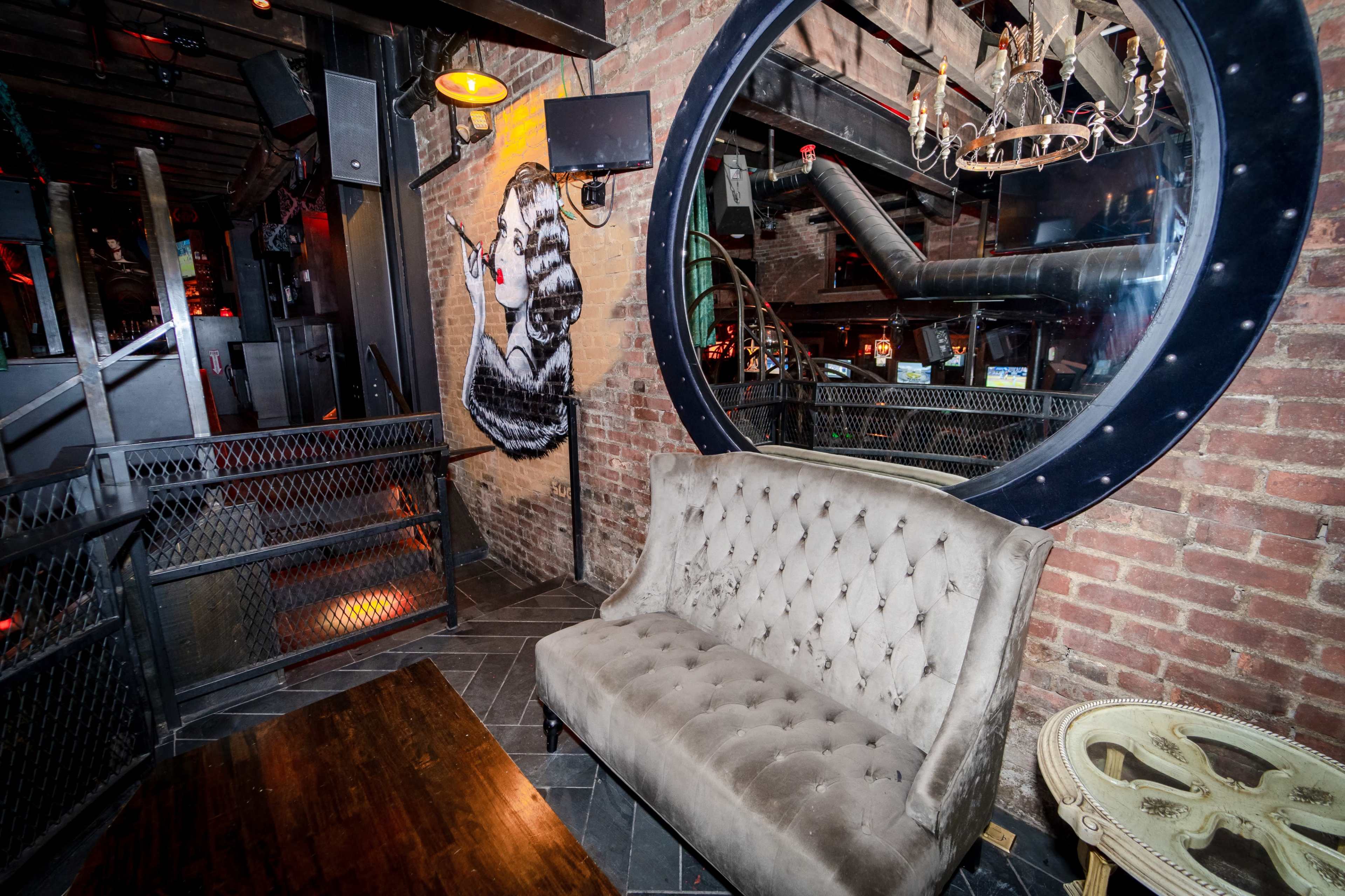 A stylish corner of a bar featuring a tufted sofa, a large round mirror, and a mural of a woman on a brick wall.
