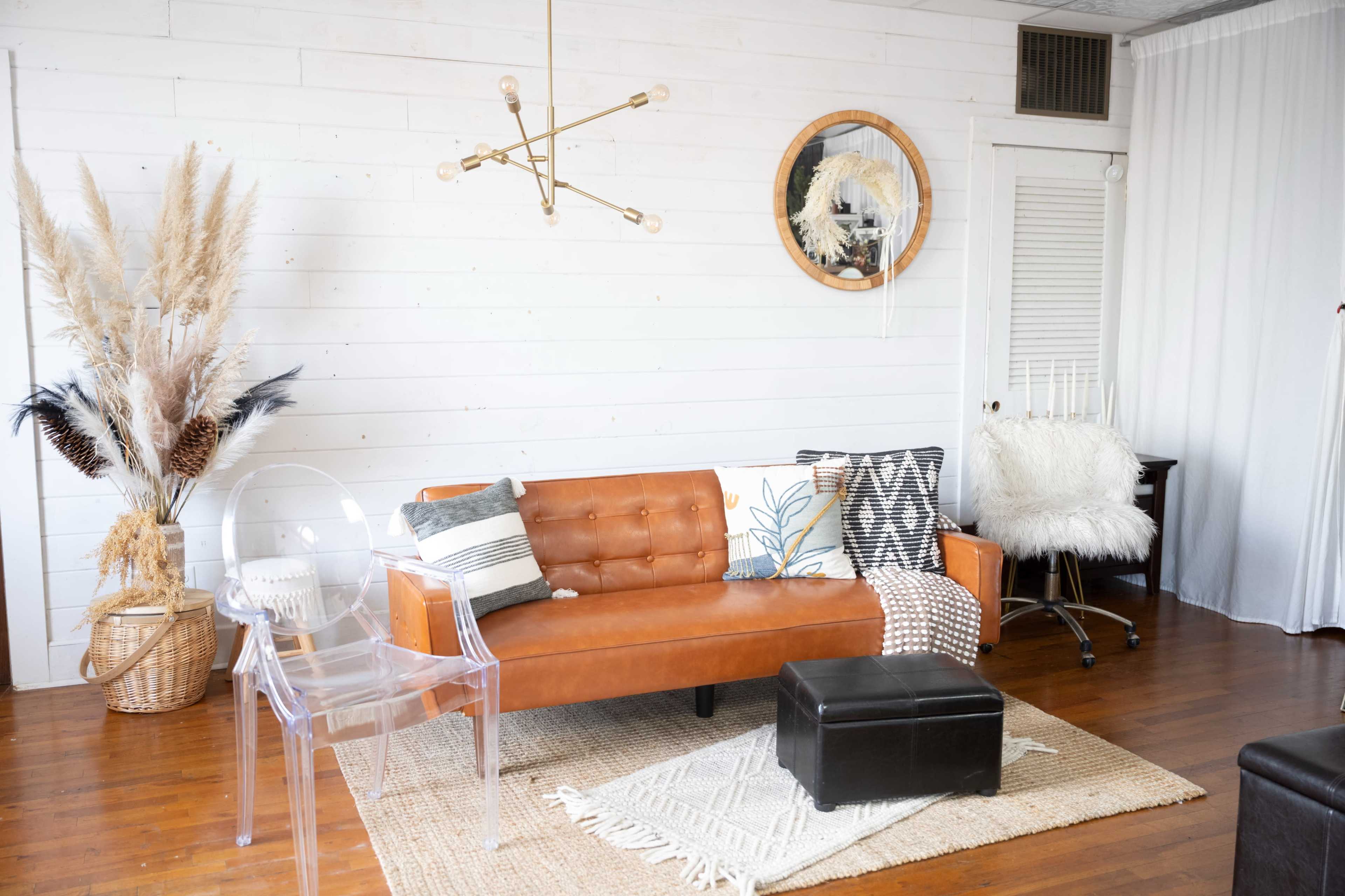 The image shows a modern living room with a brown leather sofa, decorative pillows, a clear acrylic chair, and a large woven basket containing dried plants, all set against a white shiplap wall.