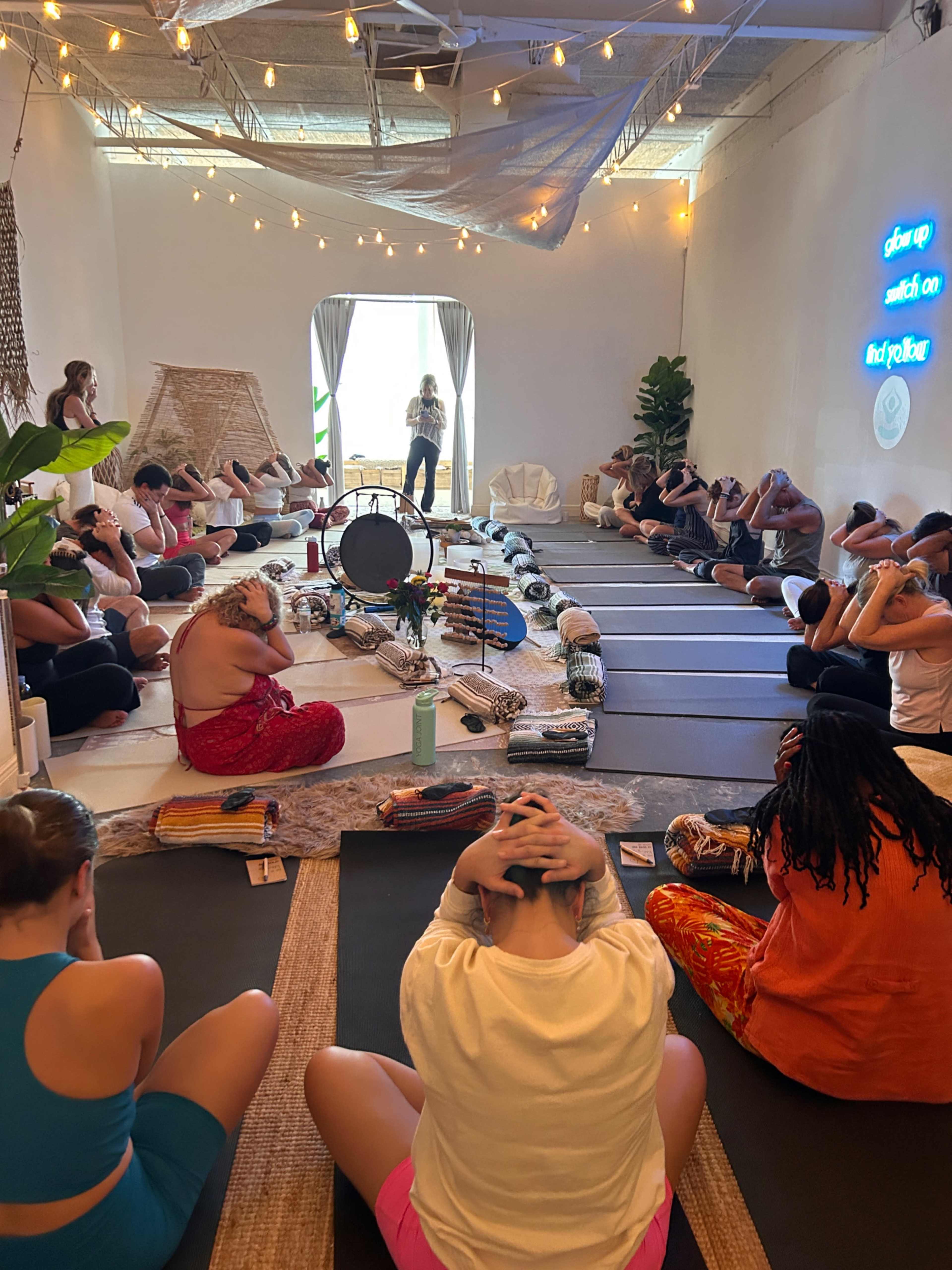 A group of individuals sits in a yoga studio, participating in a guided session with mats laid out and various instruments arranged in the center.