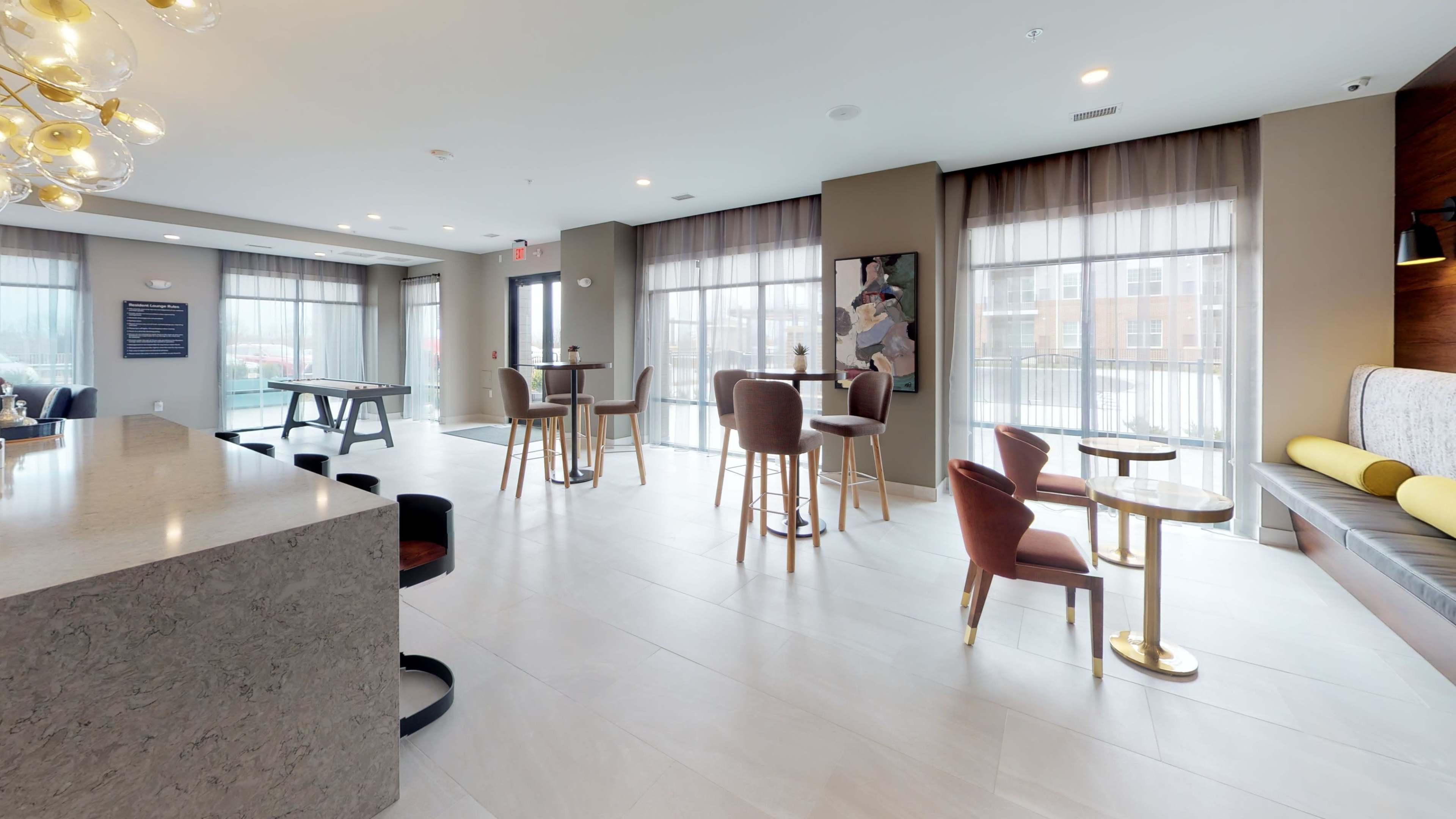 The image shows a modern gathering area with tables, chairs, and large windows allowing natural light to fill the space.