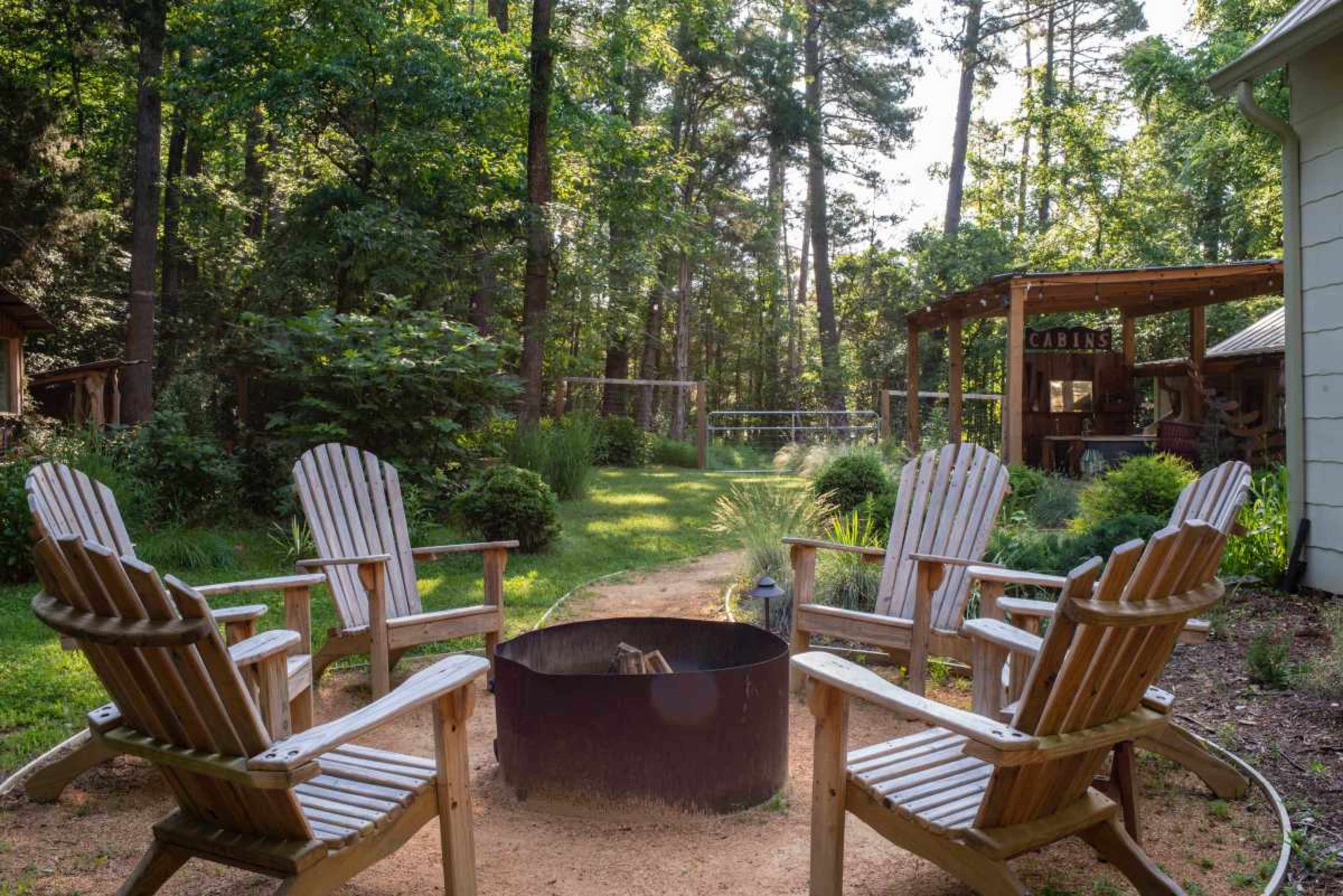 A fire pit surrounded by wooden chairs in a wooded area with green grass and trees.