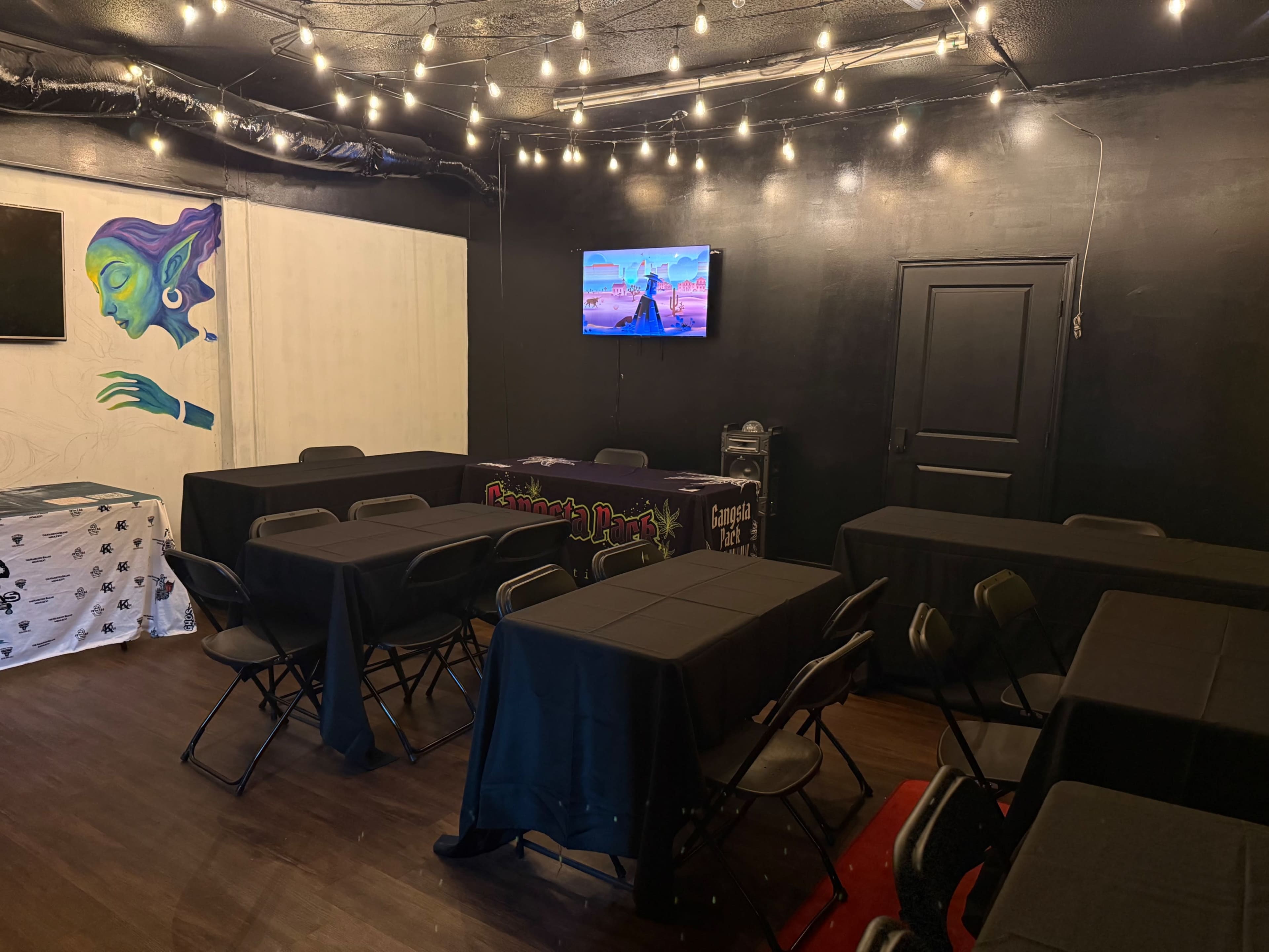 The image shows a well-lit room with several black tables and chairs, featuring a television on one wall and artwork on another.
