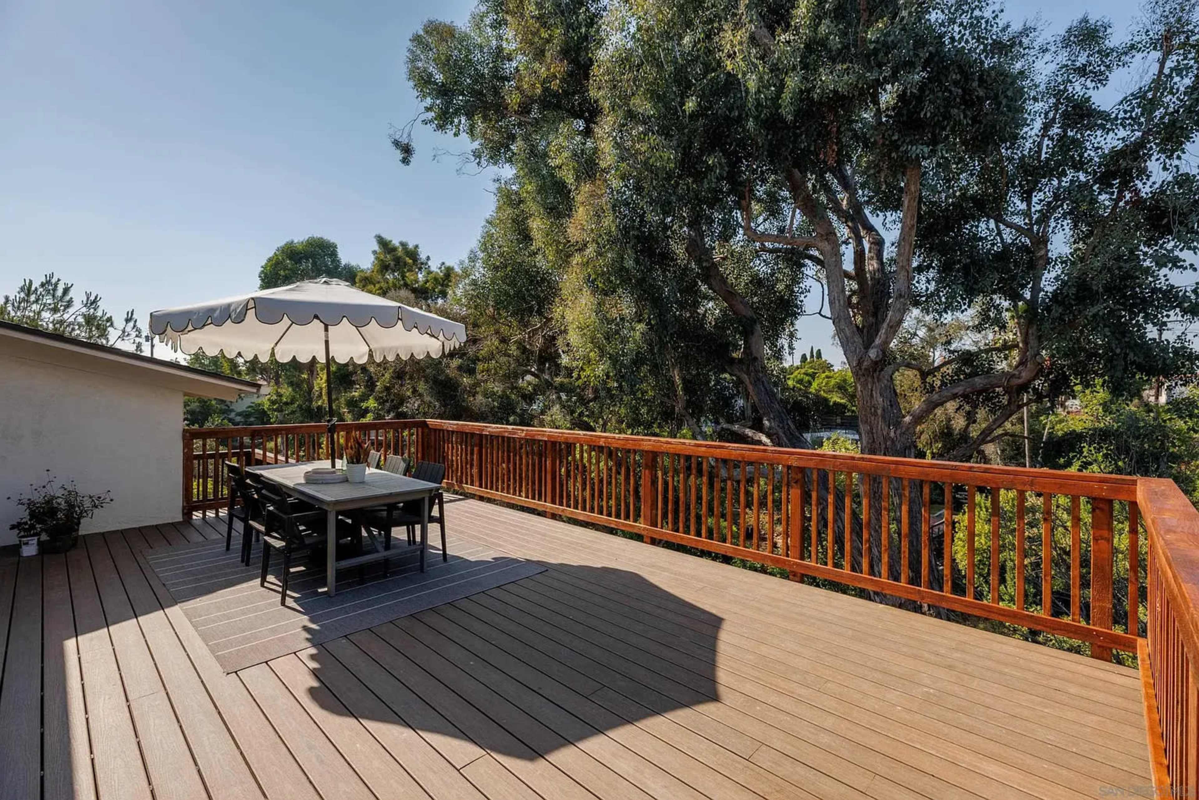 The image shows a wooden deck with a dining table and chairs under a large umbrella, surrounded by trees.