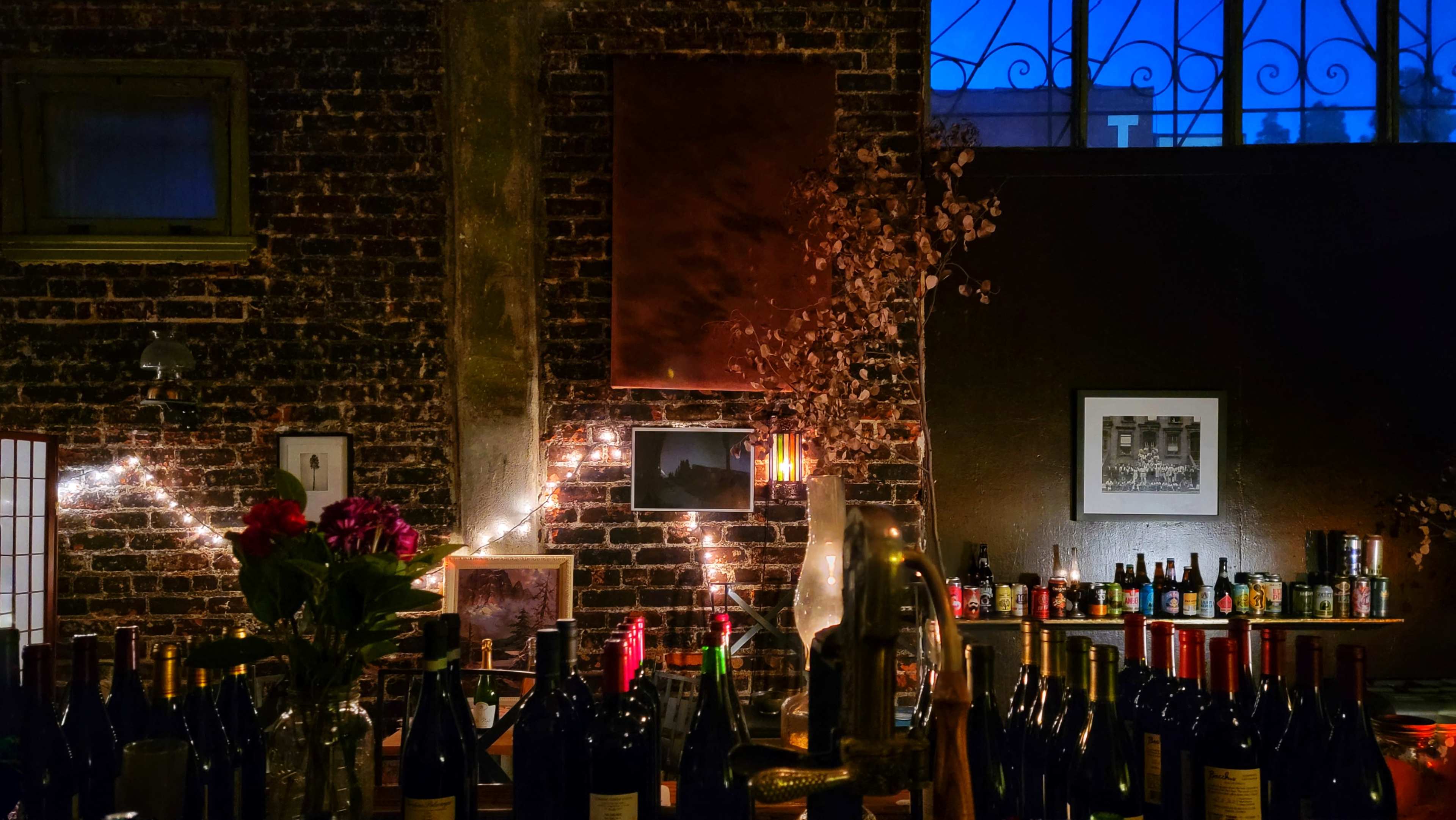 A dimly lit bar features a variety of wine bottles in the foreground, with exposed brick walls and decorative lighting creating a cozy atmosphere.