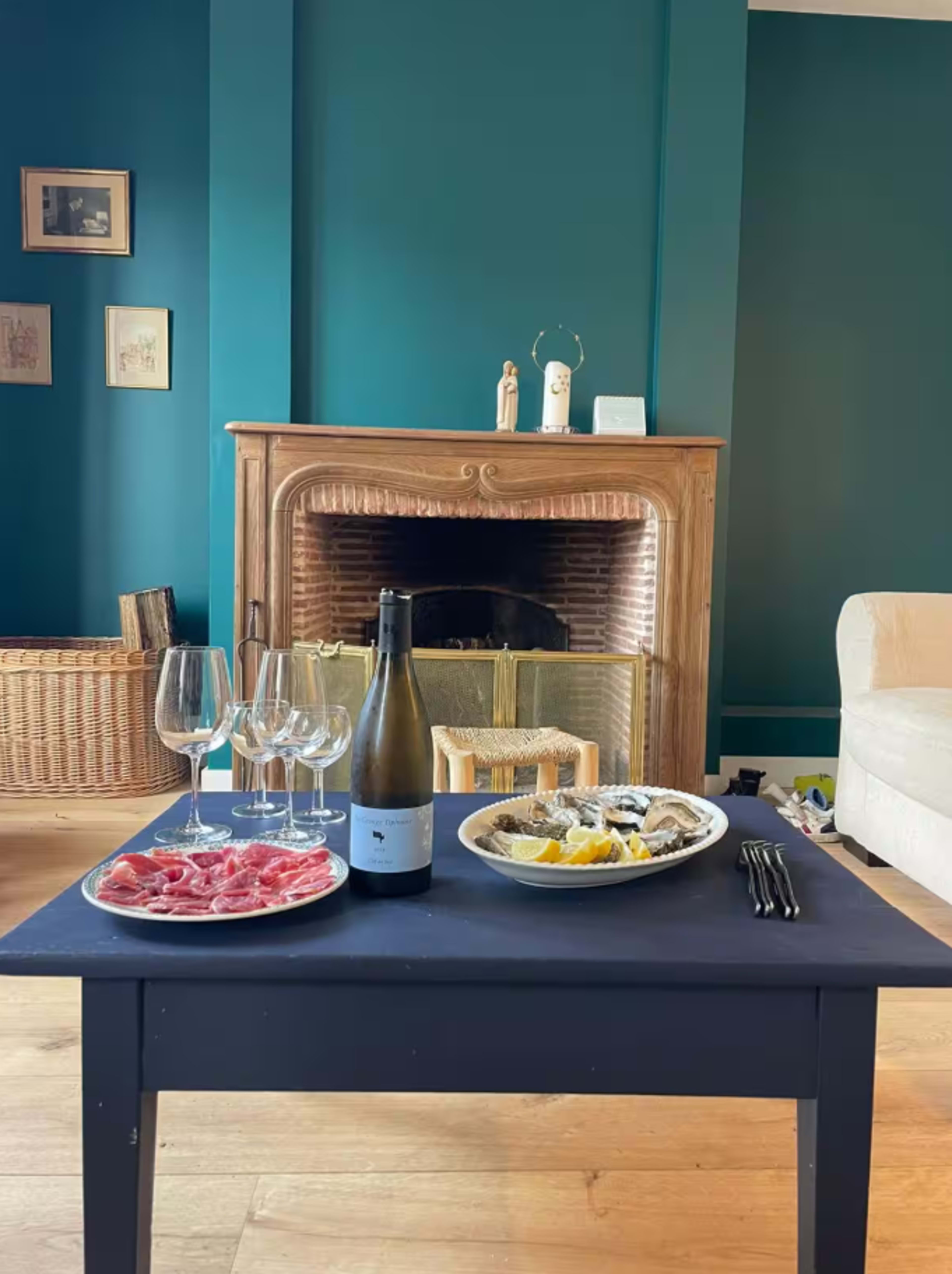 A table with a plate of sliced meat, a platter of oysters, and a bottle of wine sits in front of a fireplace in a room with teal walls.