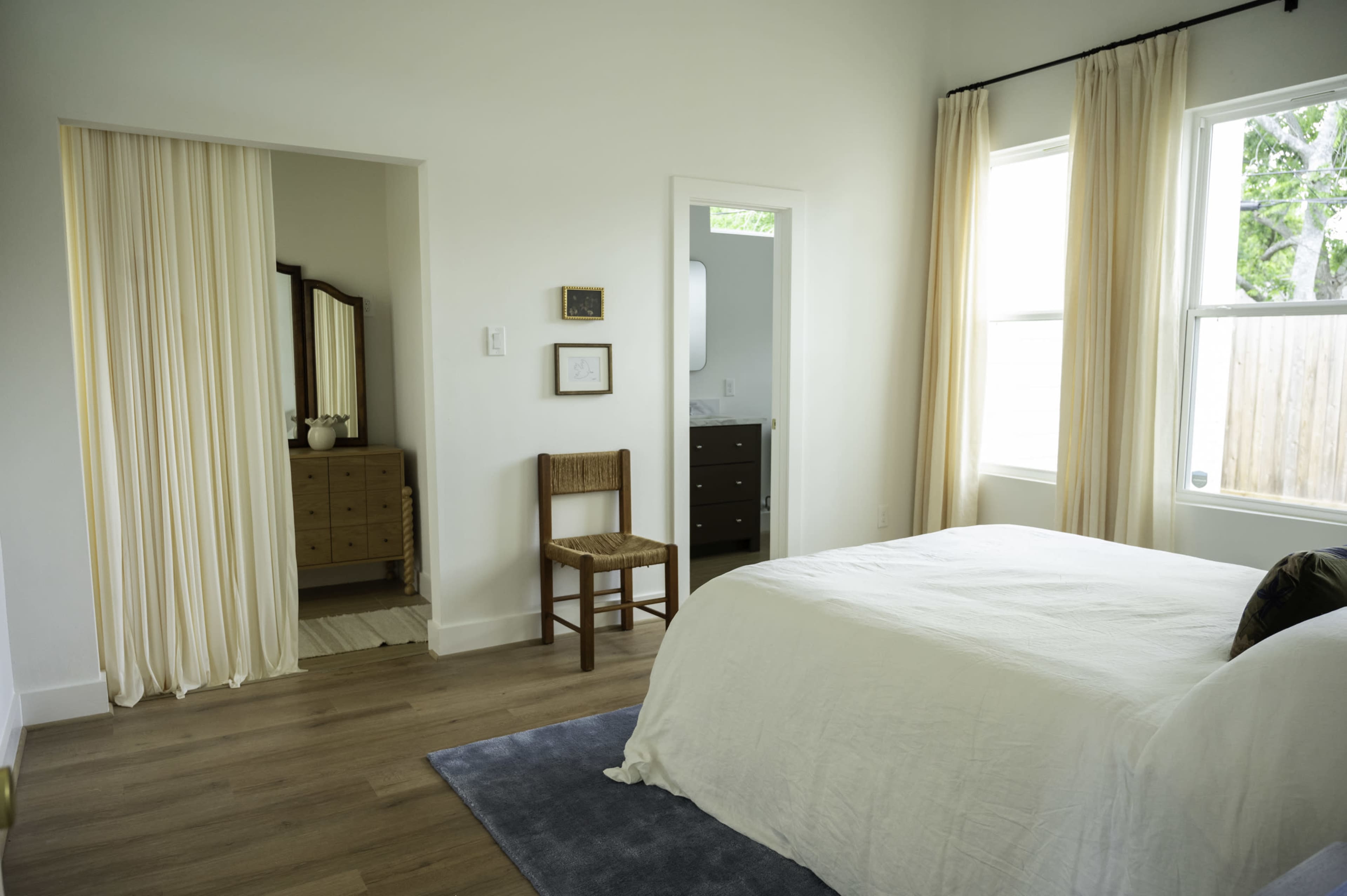 The image shows a bright, minimalist bedroom featuring a bed with white bedding, a wooden chair, and a dresser visible through an open doorway.