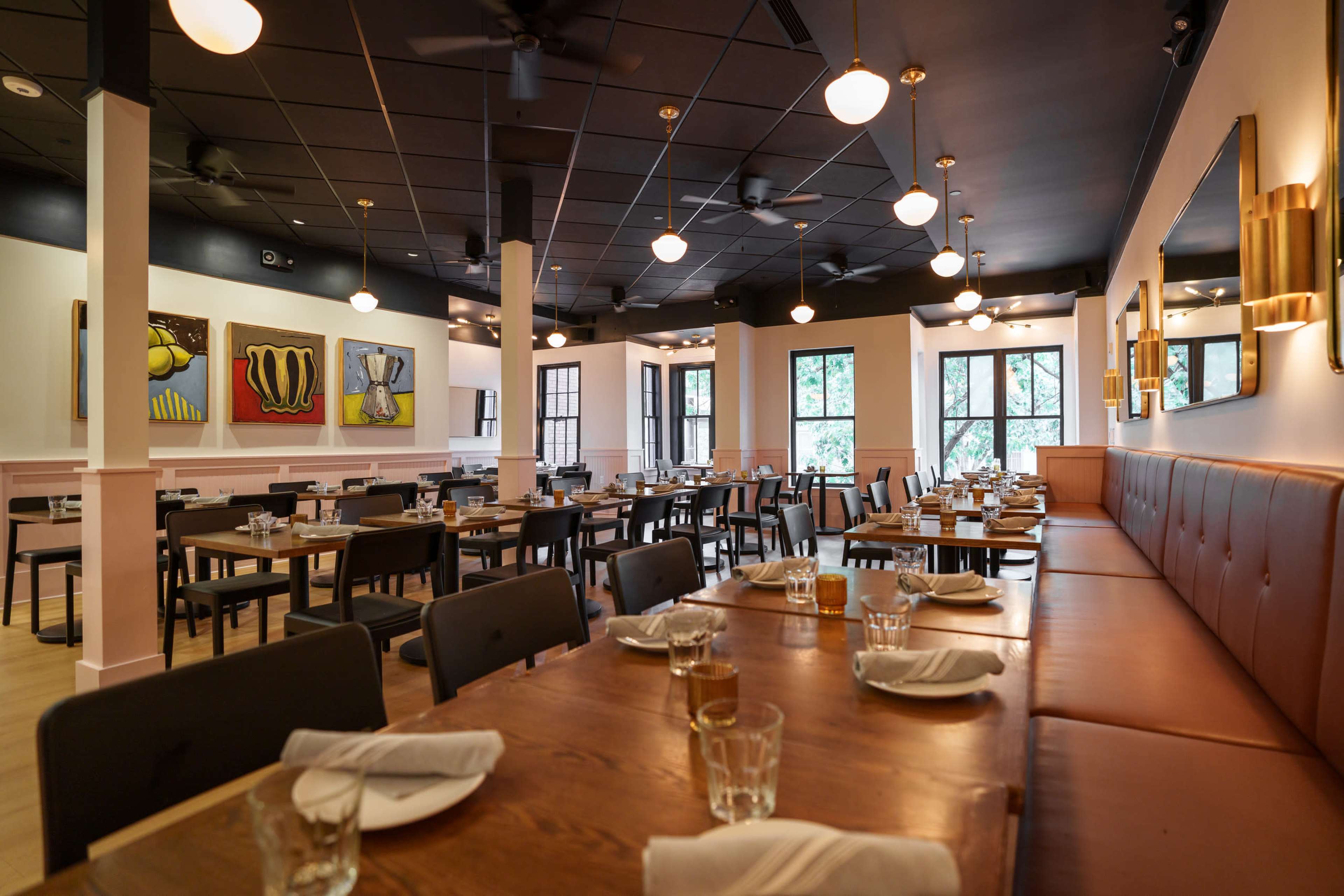 The image shows a modern restaurant interior with wooden tables and brown leather seating, illuminated by pendant lights and featuring colorful artwork on the walls.