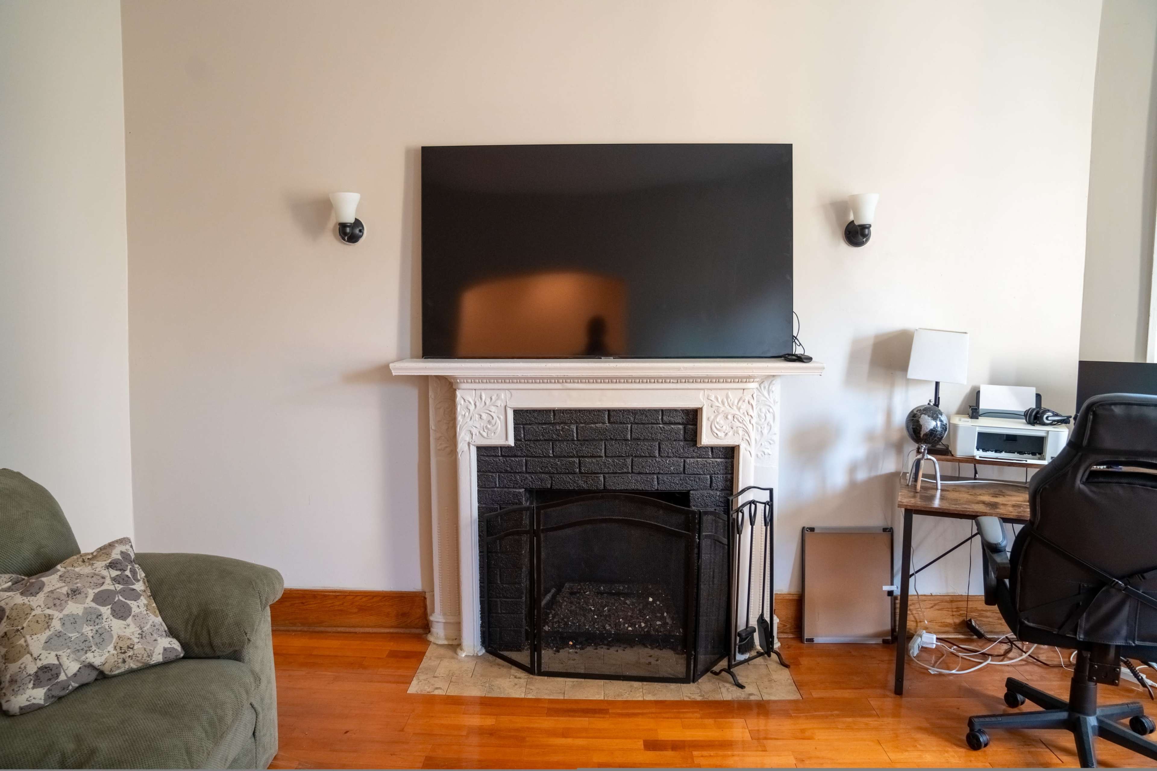 The image shows a living room with a black fireplace, a large flat-screen television mounted above it, a green sofa on the left, and a wooden desk with a computer on the right.