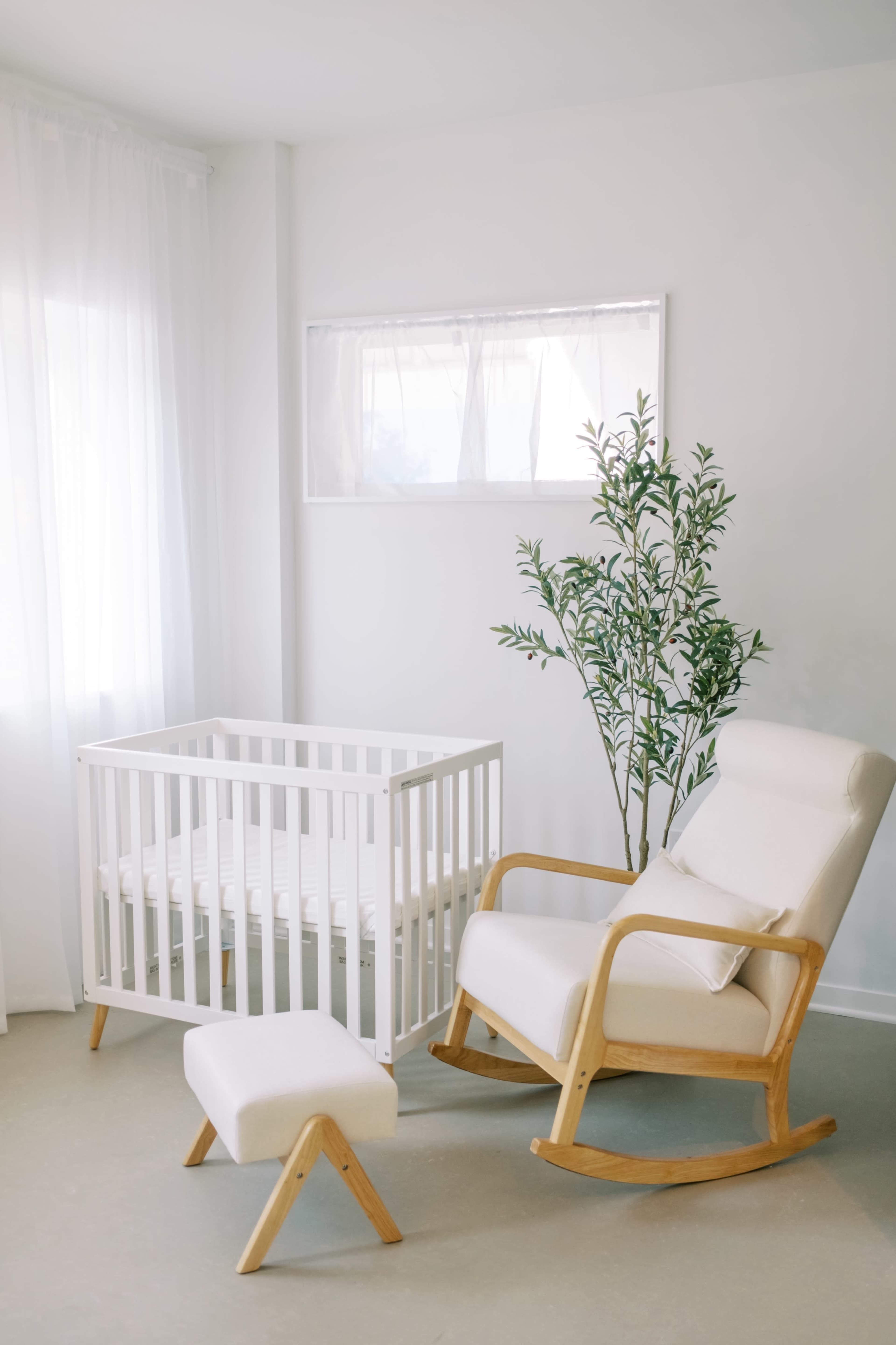 The image shows a bright nursery featuring a white crib, a rocking chair, a footrest, and a potted plant near a large window.
