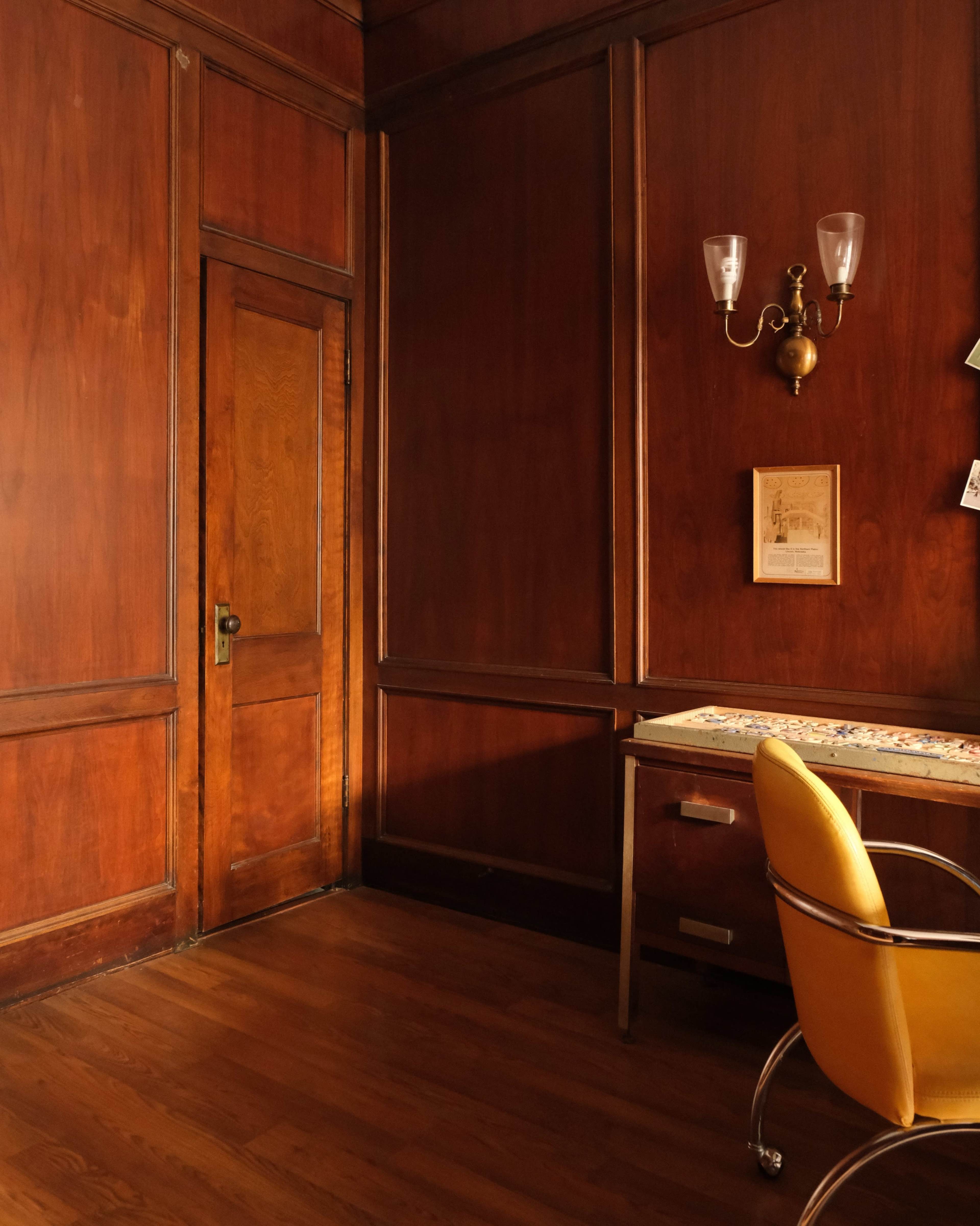 The image shows a room with wooden walls, a door on the left, a desk with a patterned surface, and an orange chair positioned in front of it.
