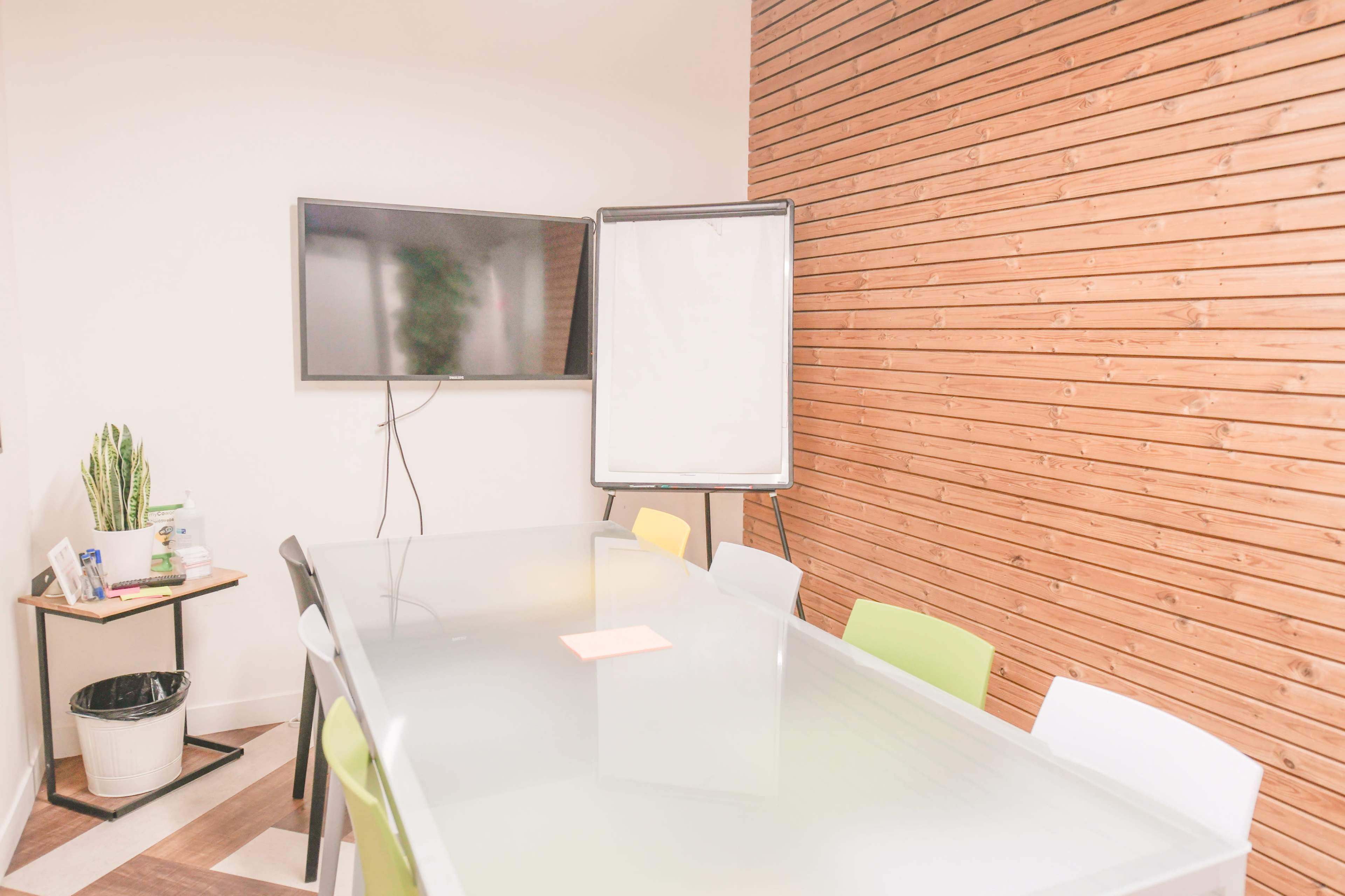 A modern conference room with a glass table, several colorful chairs, a TV mounted on the wall, and a whiteboard on an easel.