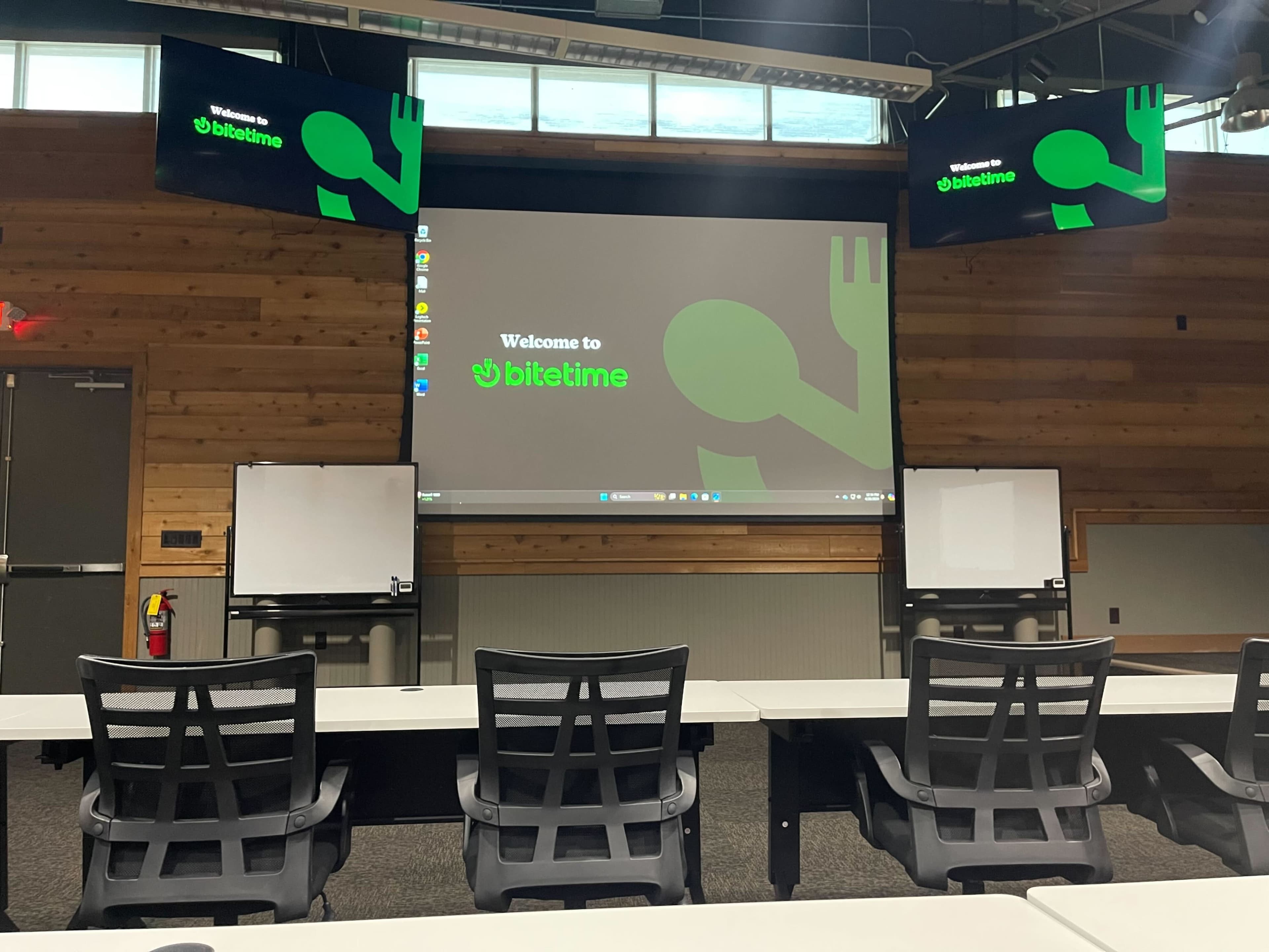 The image shows a conference room with a large screen displaying "Welcome to bitetime" and two additional monitors on either side, along with empty tables and chairs set up for a meeting.