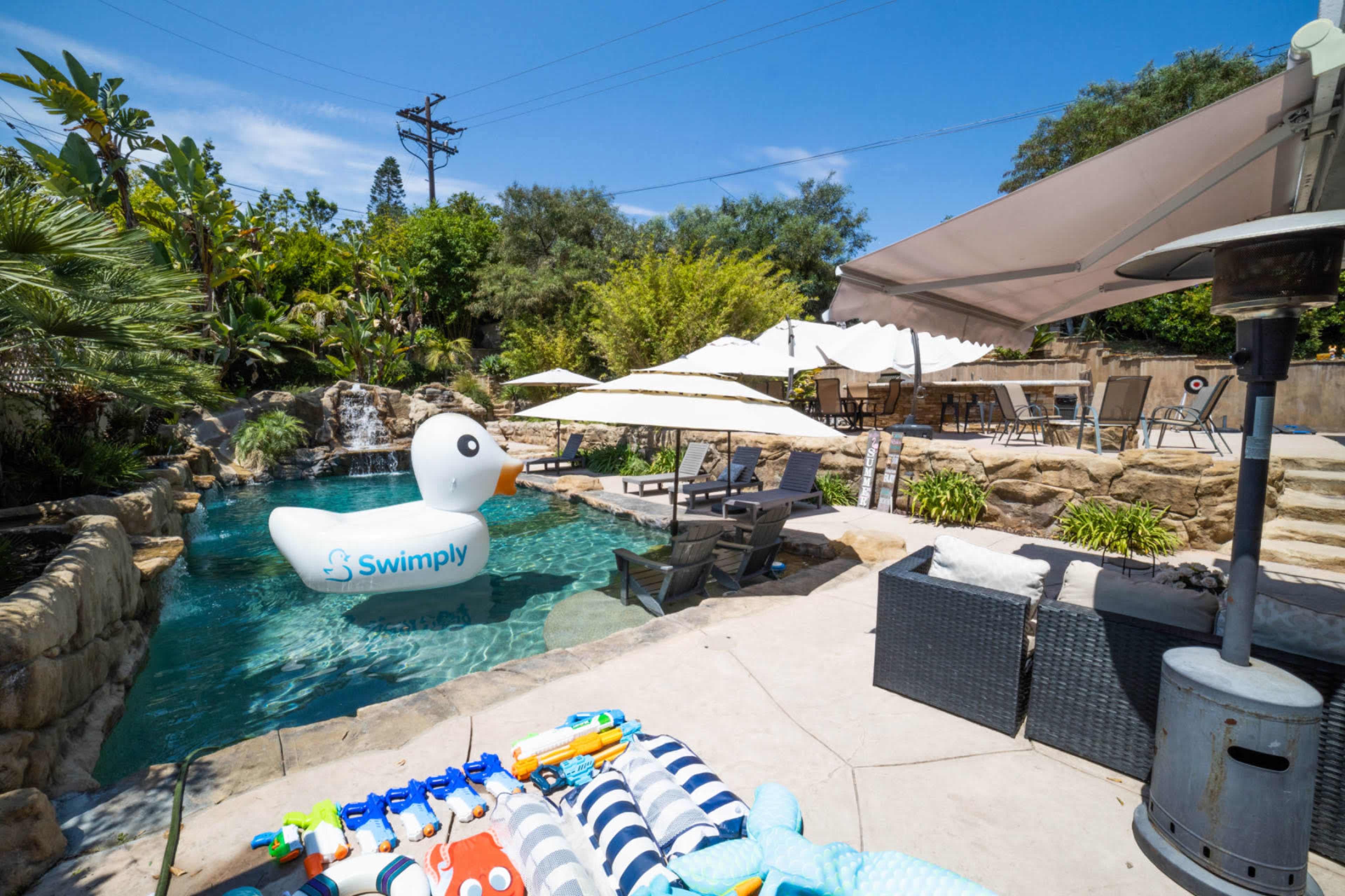 A large inflatable duck floats in a residential pool surrounded by lounge chairs and shaded seating areas.