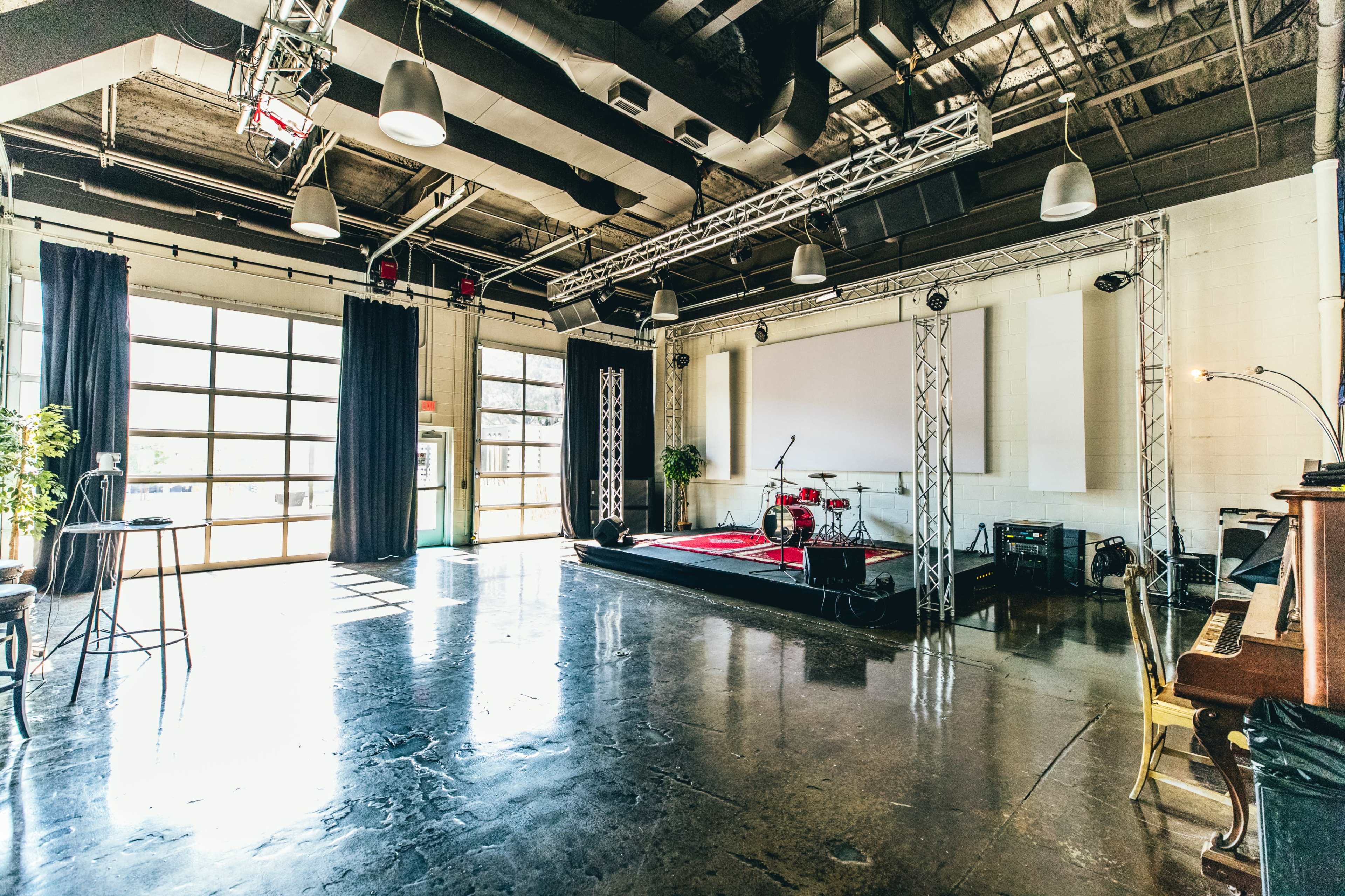 The image shows a spacious indoor venue with a stage equipped for performances, featuring a drum set, lighting rigs, and large windows allowing natural light to enter.