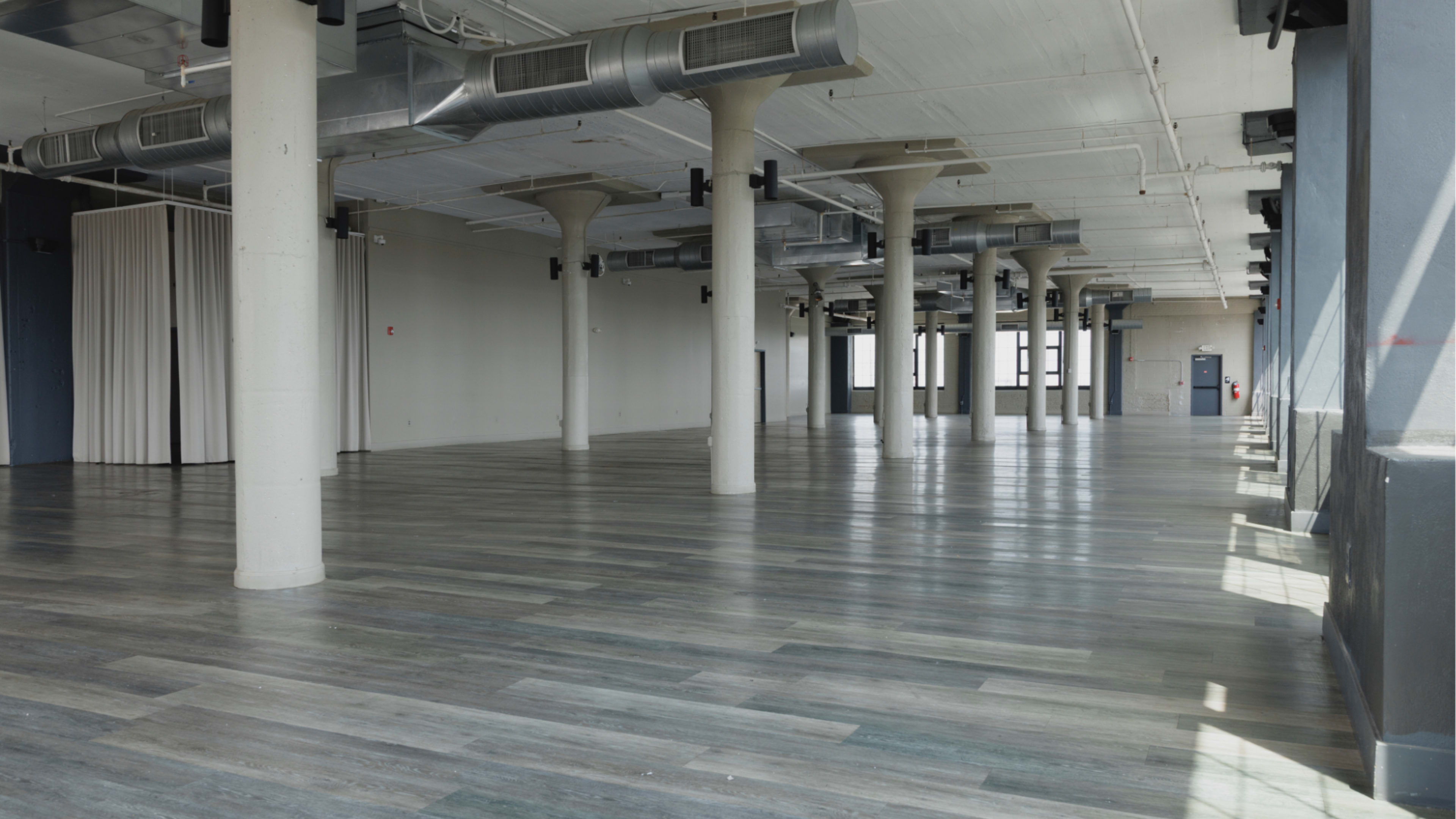 The image shows a spacious, empty room with wooden flooring and several support columns, illuminated by natural light coming through large windows.
