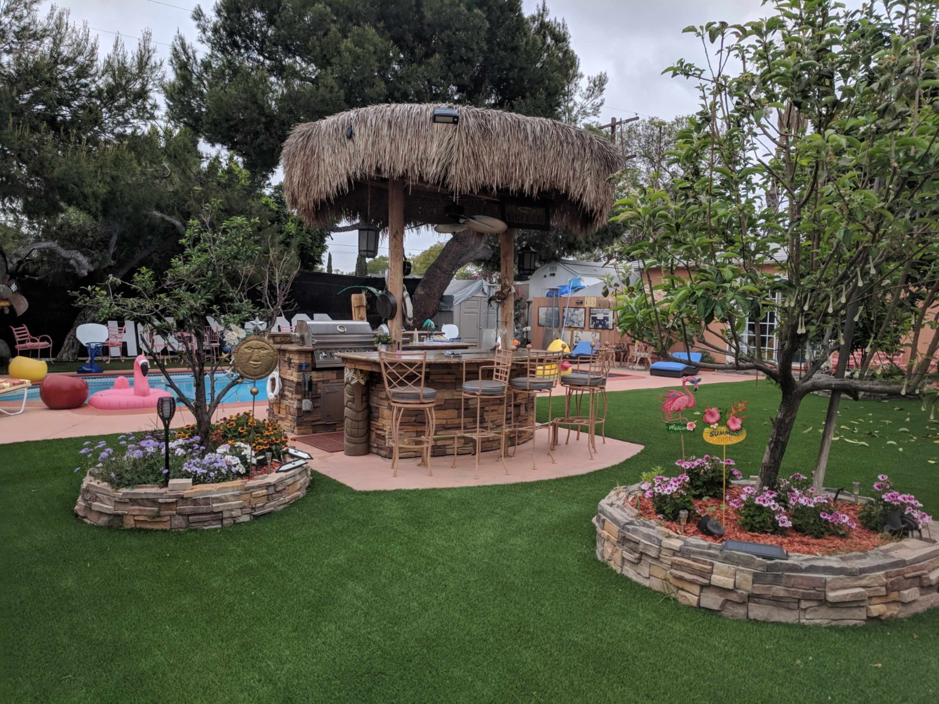 A backyard with a thatched-roof bar surrounded by colorful flowers and greenery, featuring a swimming pool and various recreational items.