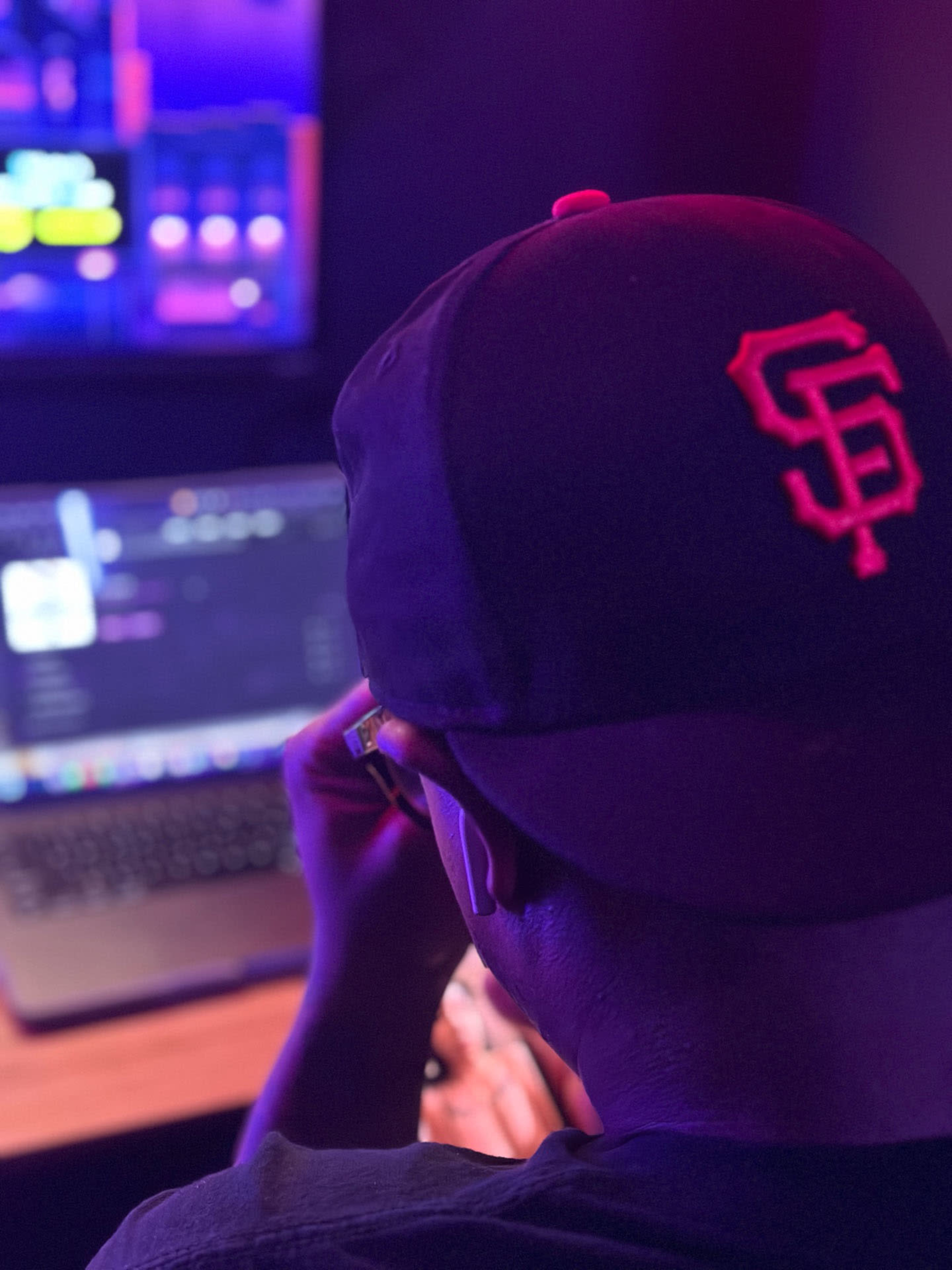 A person wearing a black hat sits in front of a laptop, illuminated by colorful lights in a dimly lit room.