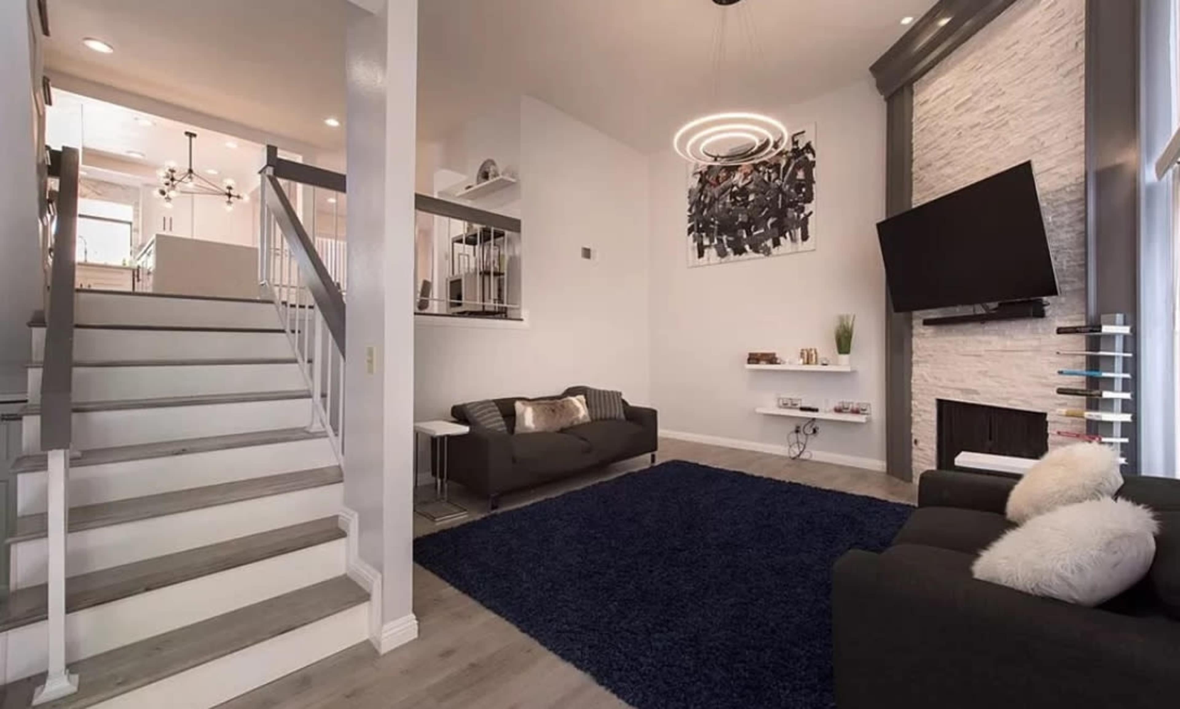 A modern living room with a staircase, a large rug, two sofas, and a wall-mounted television.