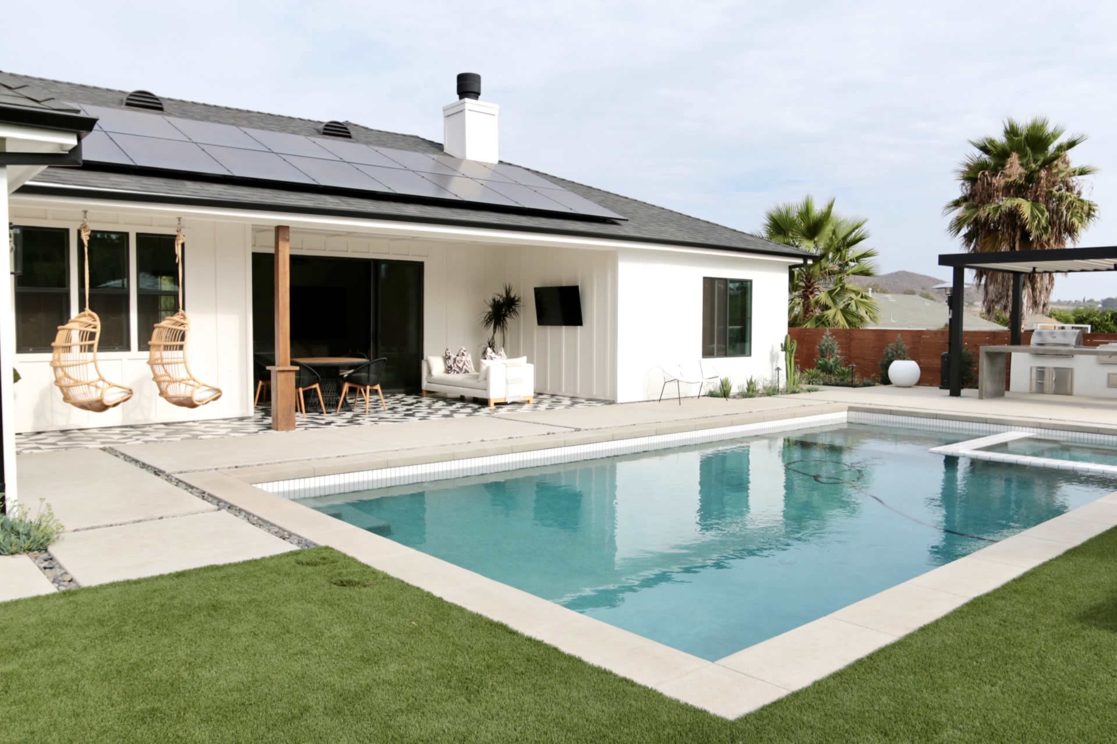 The image shows a modern backyard featuring a swimming pool in front of a single-story white house with solar panels on the roof and hanging chairs on the patio.