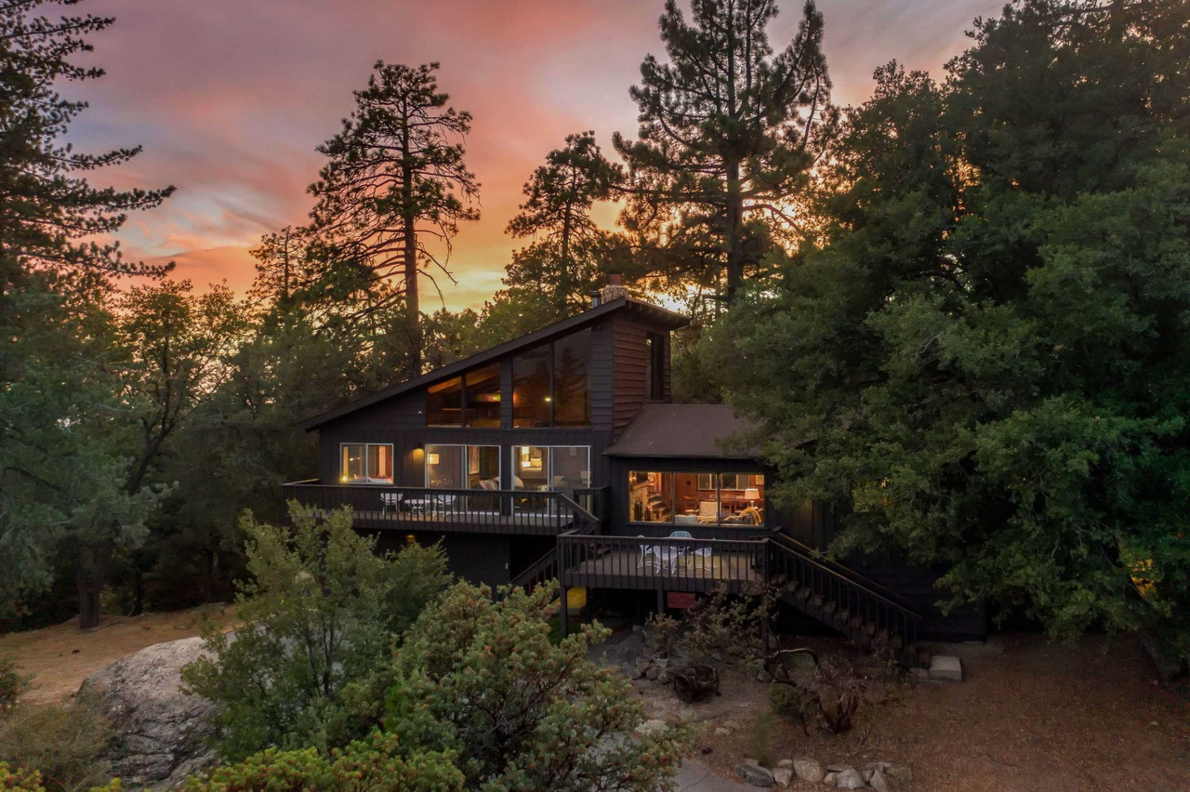 A modern cabin with large windows is surrounded by trees, set against a backdrop of a colorful sunset.