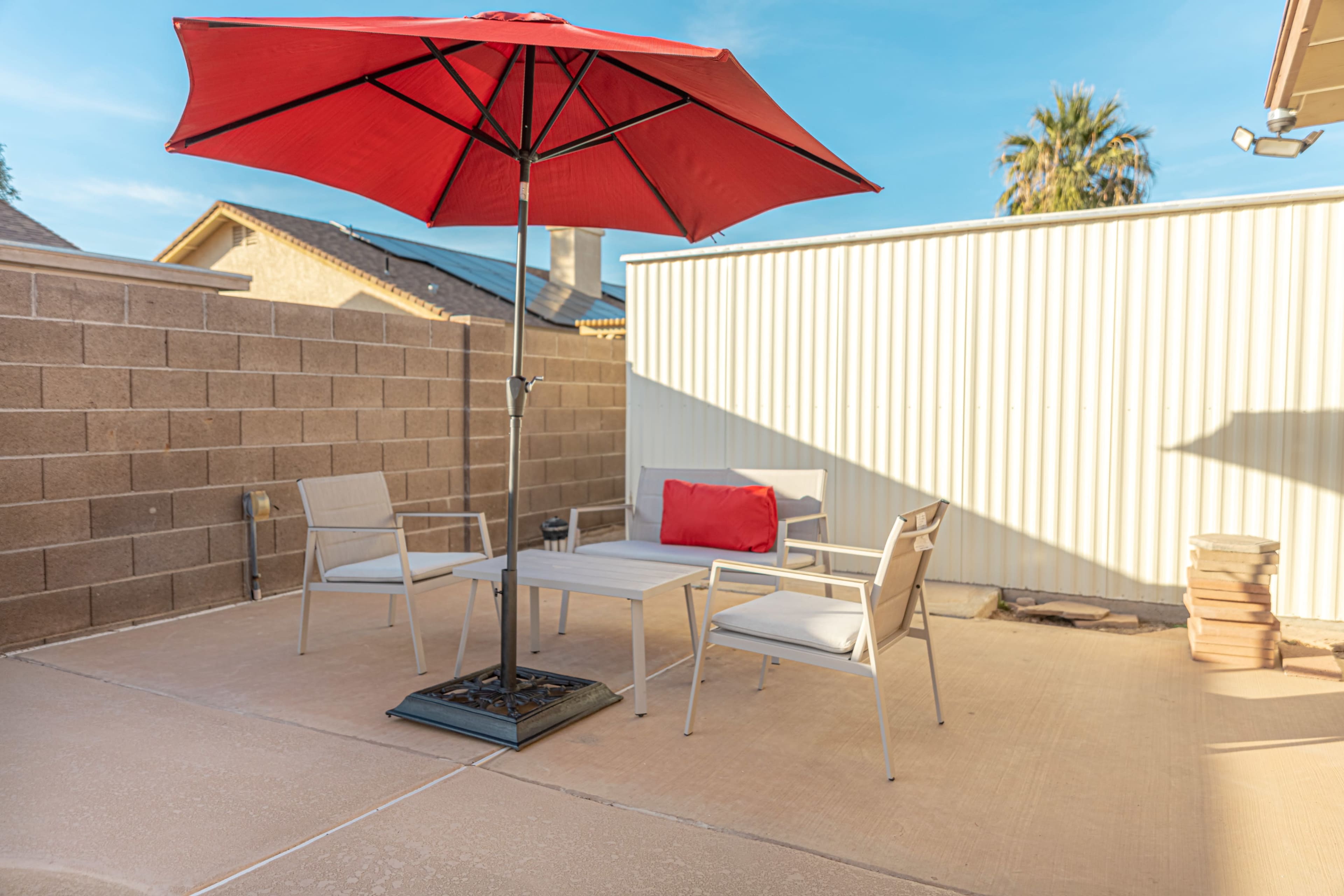 A red umbrella shades a small outdoor seating area with two chairs and a table on a concrete patio surrounded by a brick wall.