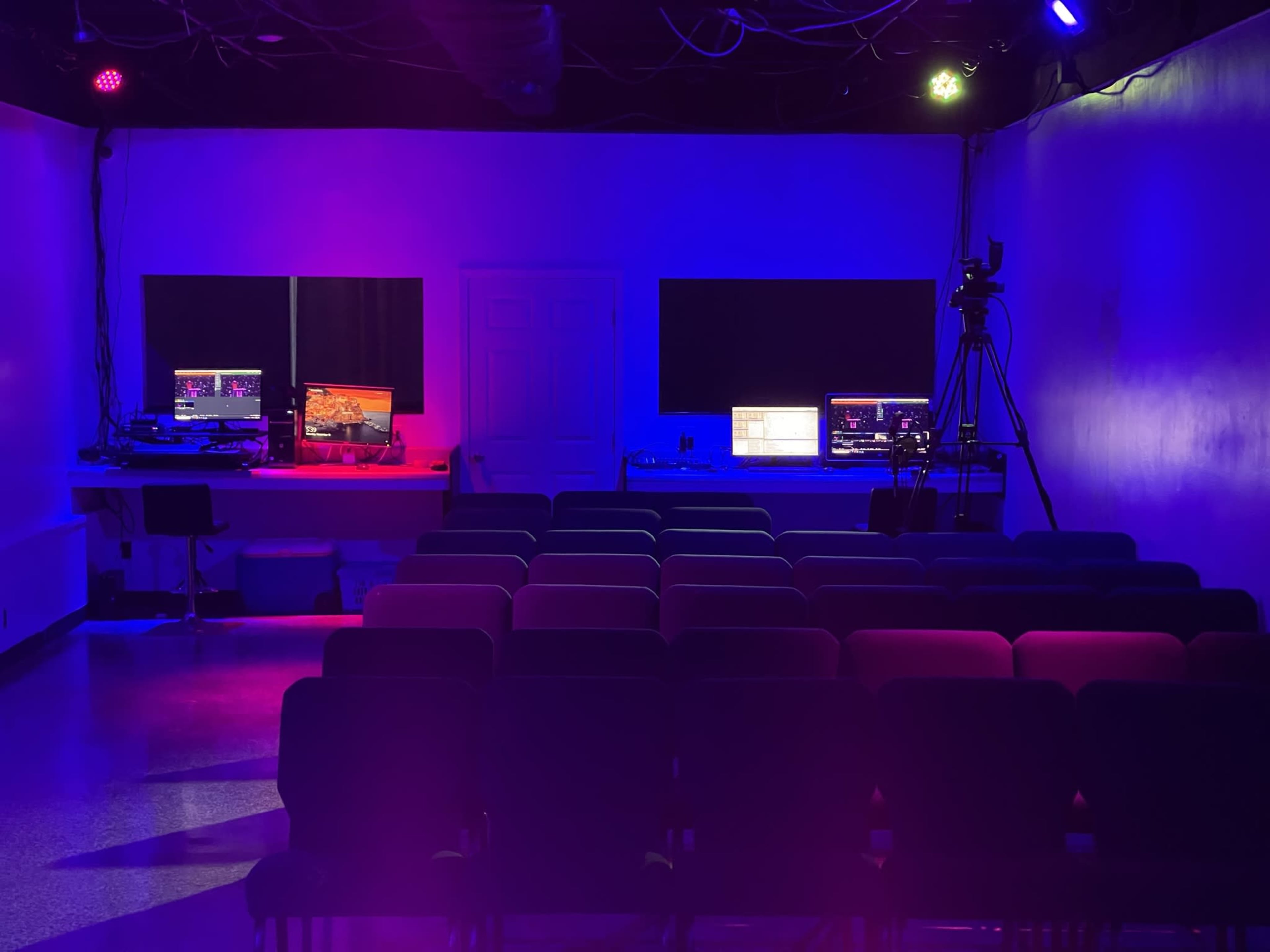 A dimly lit room with two computer setups on opposite sides and rows of chairs facing a blank wall.