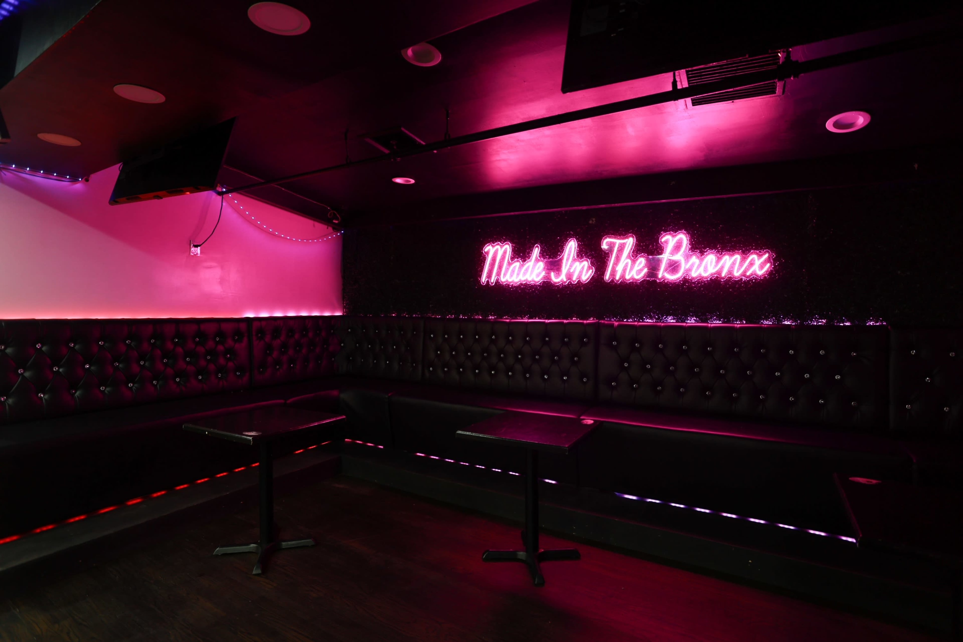 The image shows a dimly lit lounge area with a black tufted bench, small tables, and a neon sign that reads "Made In The Bronx" in pink light.