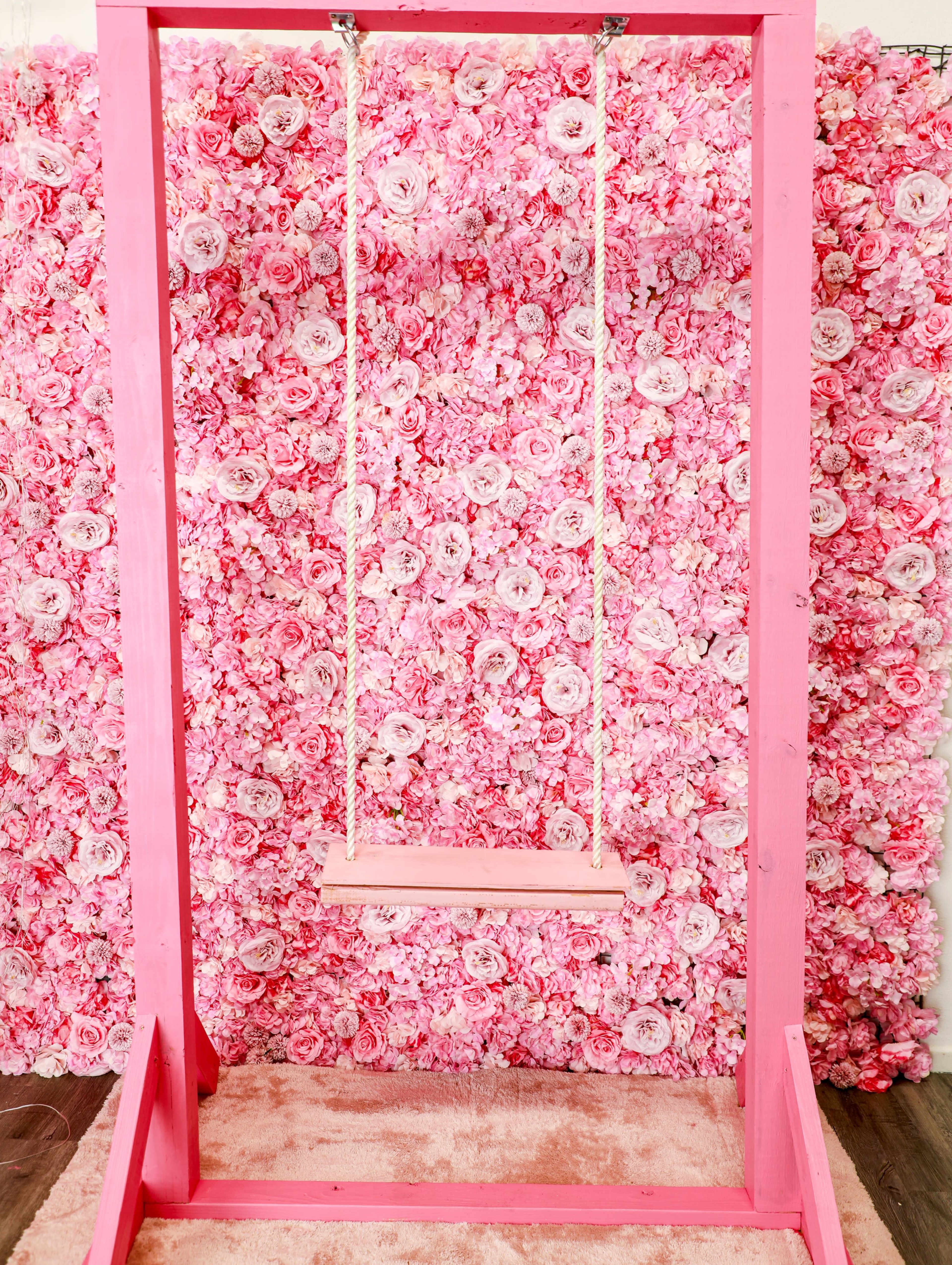 A pink swing hangs in front of a wall covered with pink floral decoration.