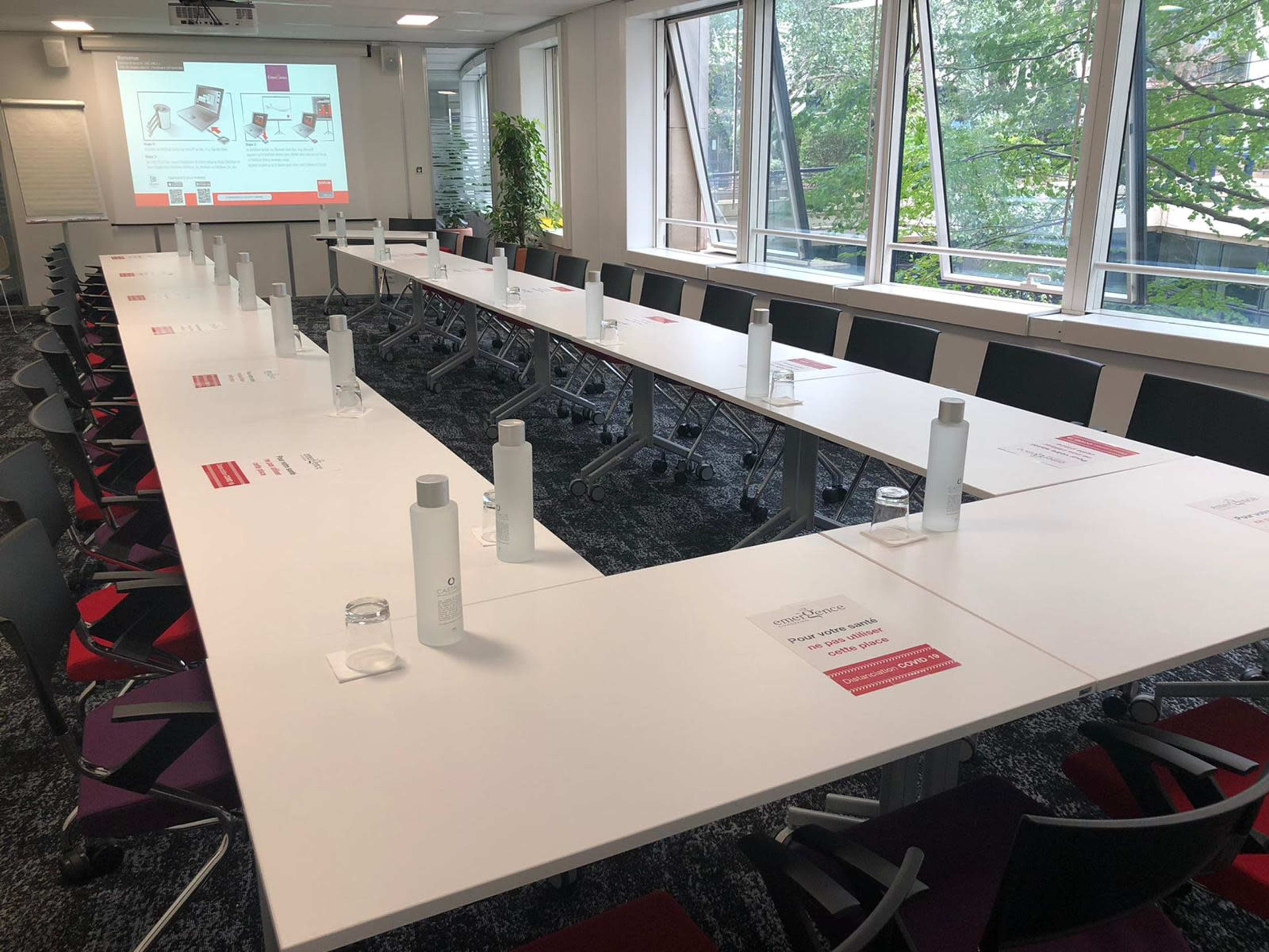 A modern conference room is set up with long tables arranged in a U-shape, featuring water bottles and printed materials, along with large windows providing a view of greenery outside.