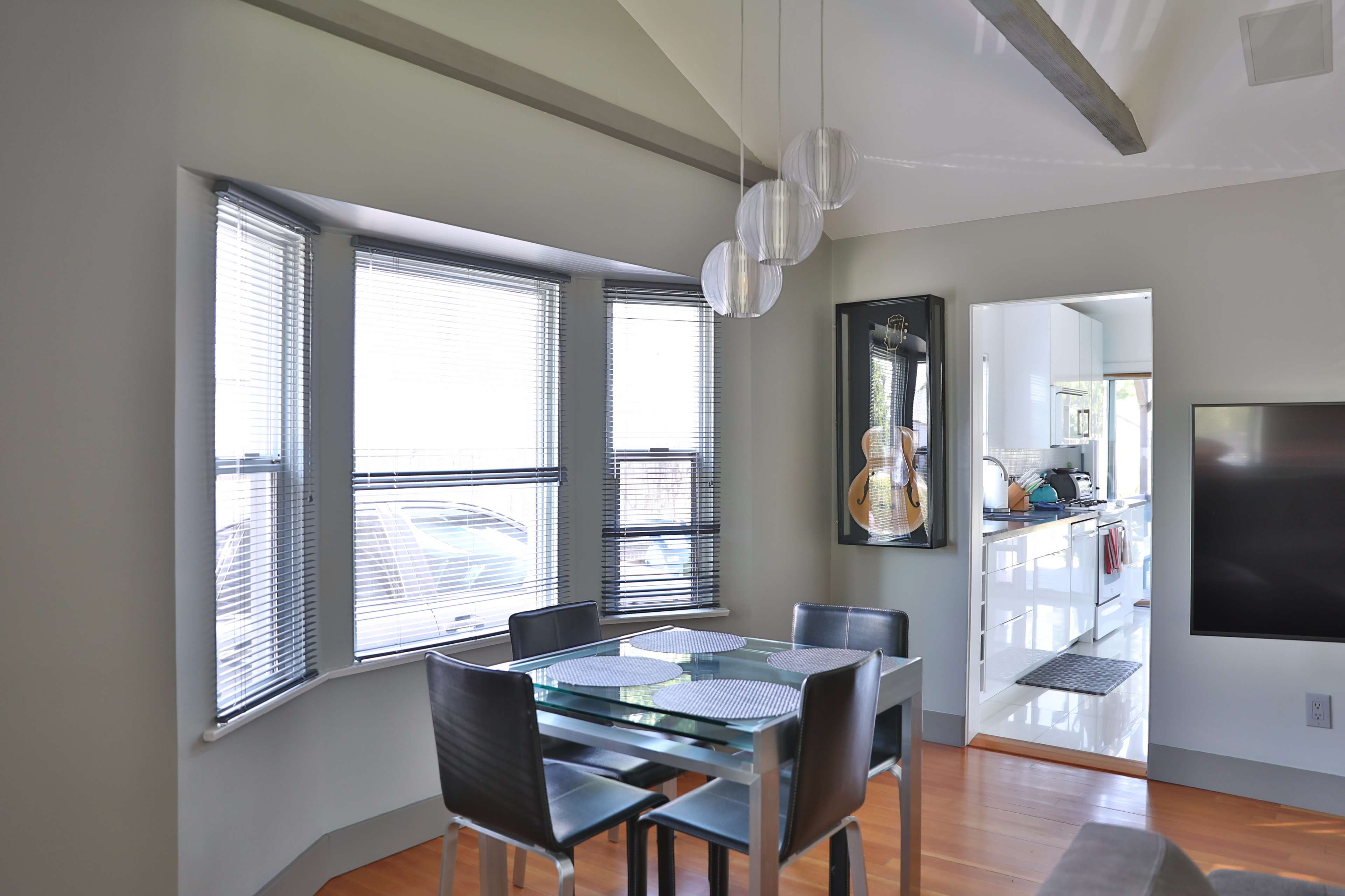 The image shows a modern dining area with a glass table and black chairs, adjacent to a well-lit kitchen through an open doorway.