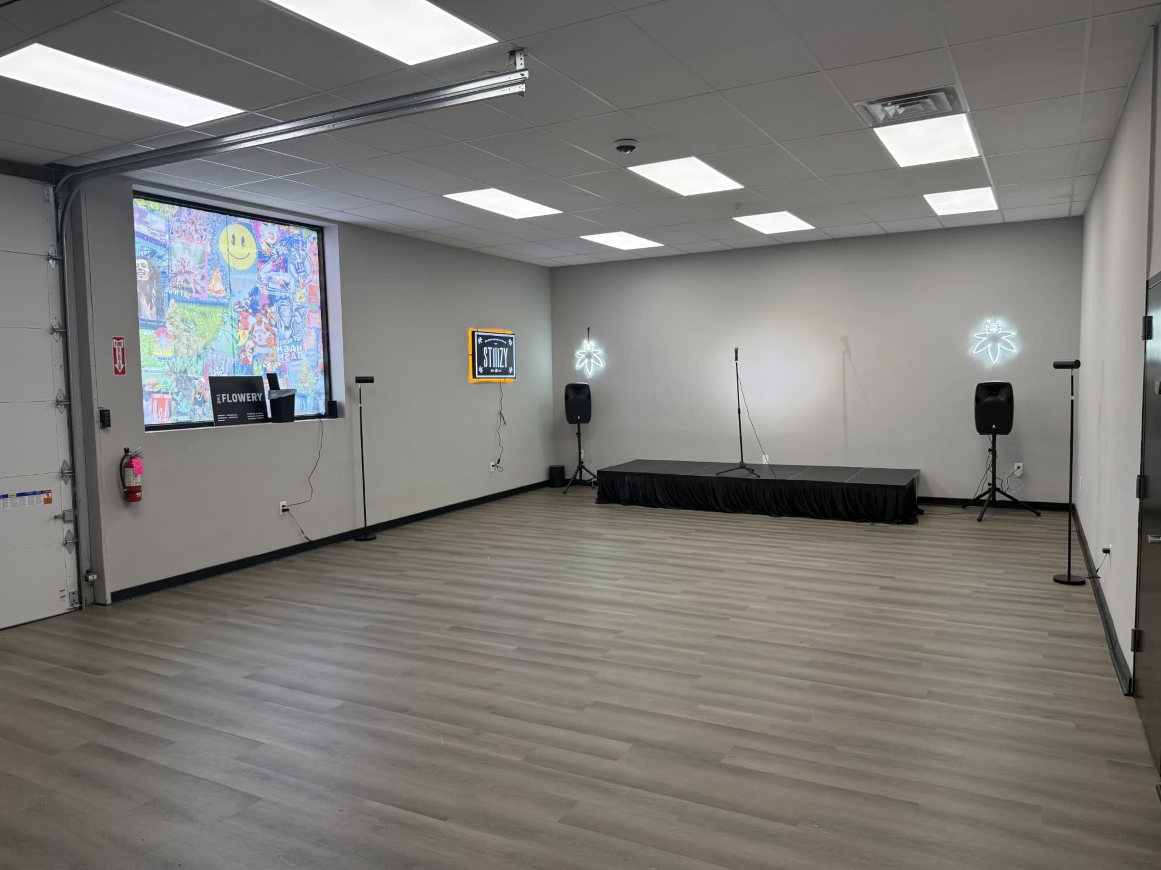 The image shows an empty event space with a small stage, two microphones on stands, colorful wall art visible through a window, and neon decorations.
