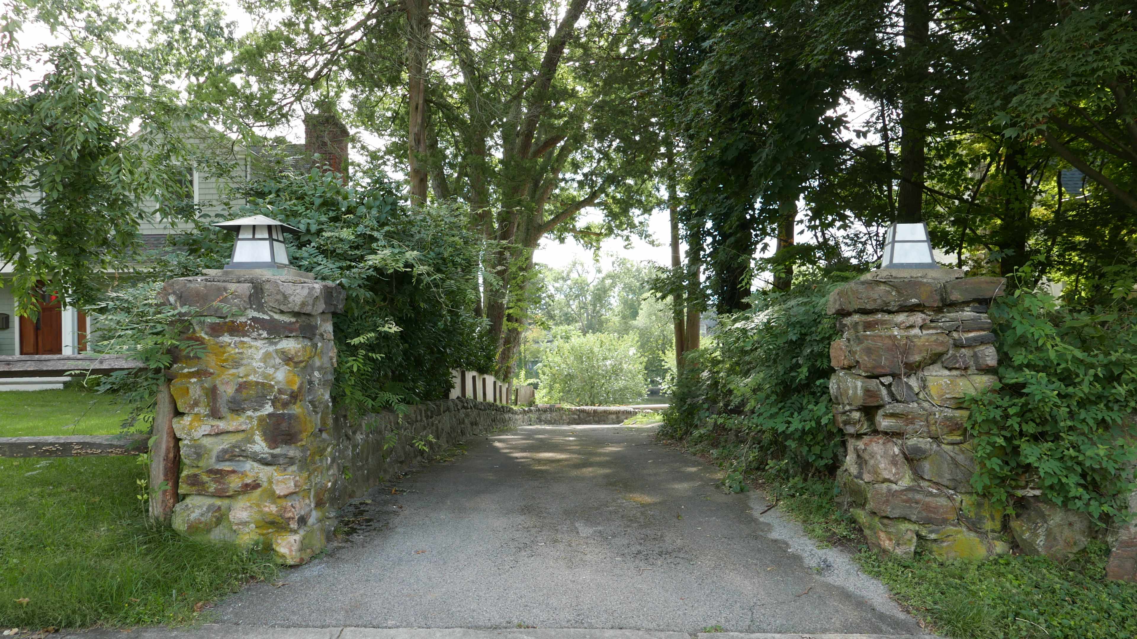 A gravel driveway lined by stone pillars leads into a wooded area flanked by trees.