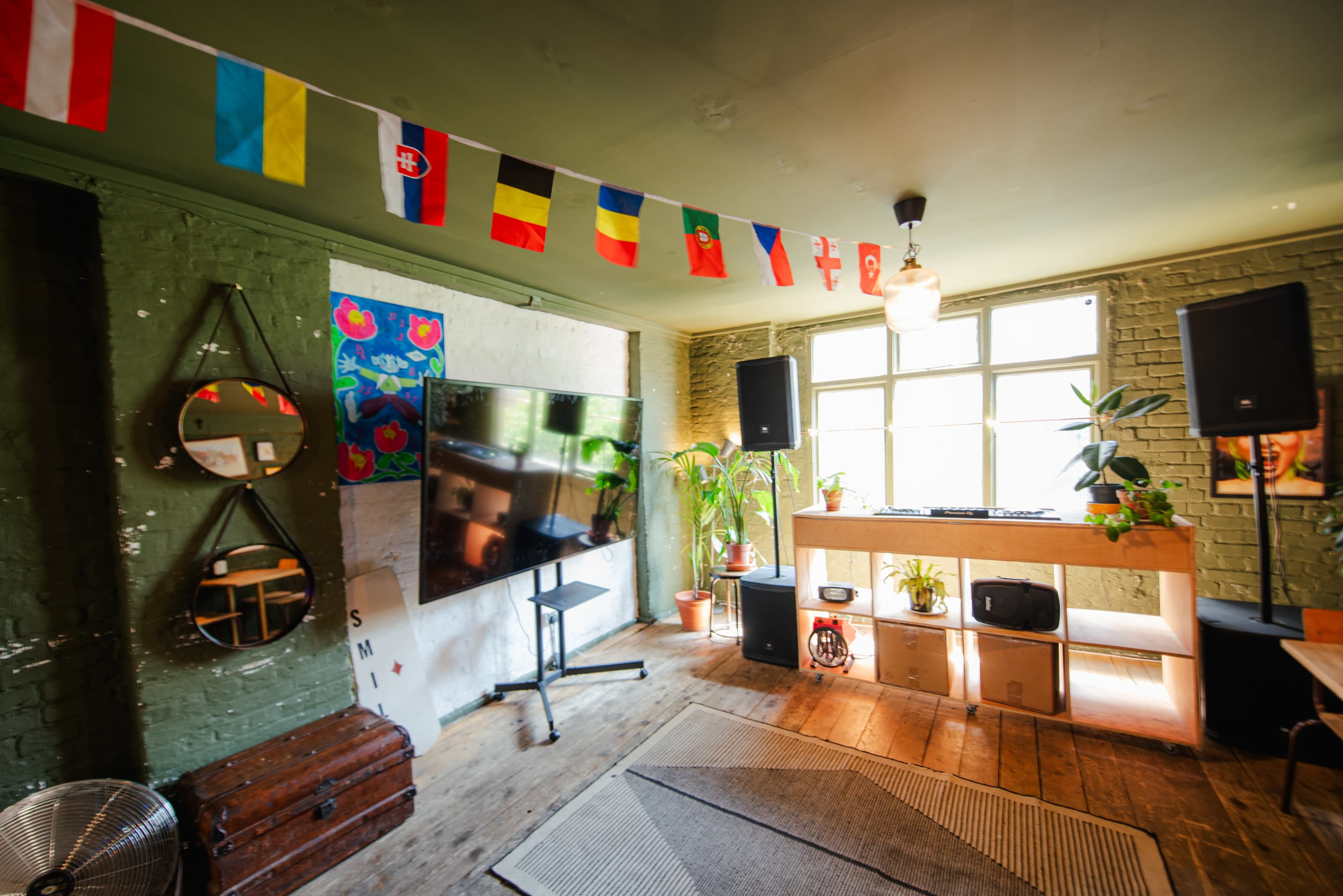 The image shows a room with a large television, speakers, and decorative flags hanging from the ceiling, alongside green-painted walls and wooden flooring.