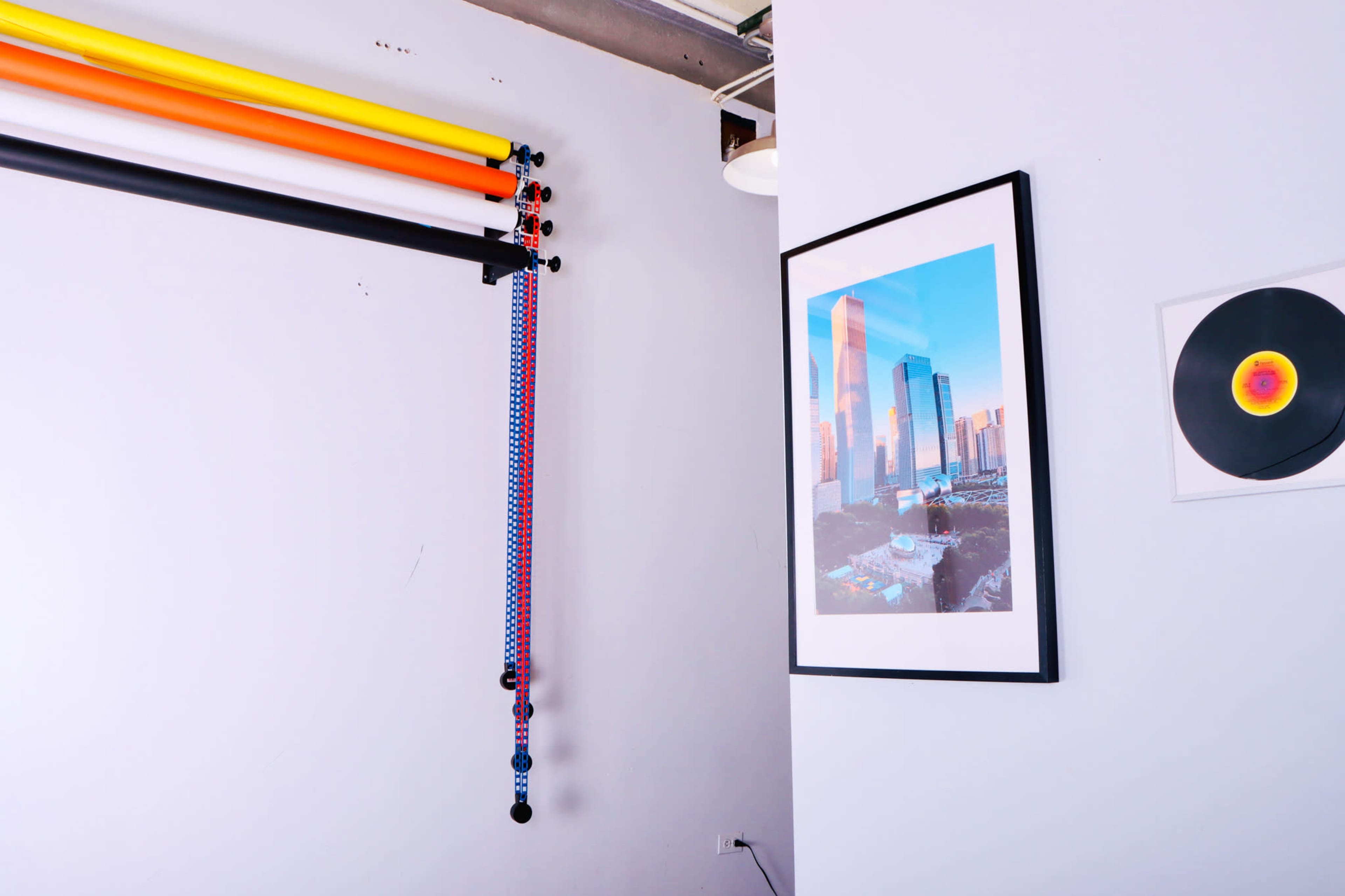 A corner of a room featuring colored backdrop rolls, a framed photograph of a cityscape, and a vinyl record on the wall.