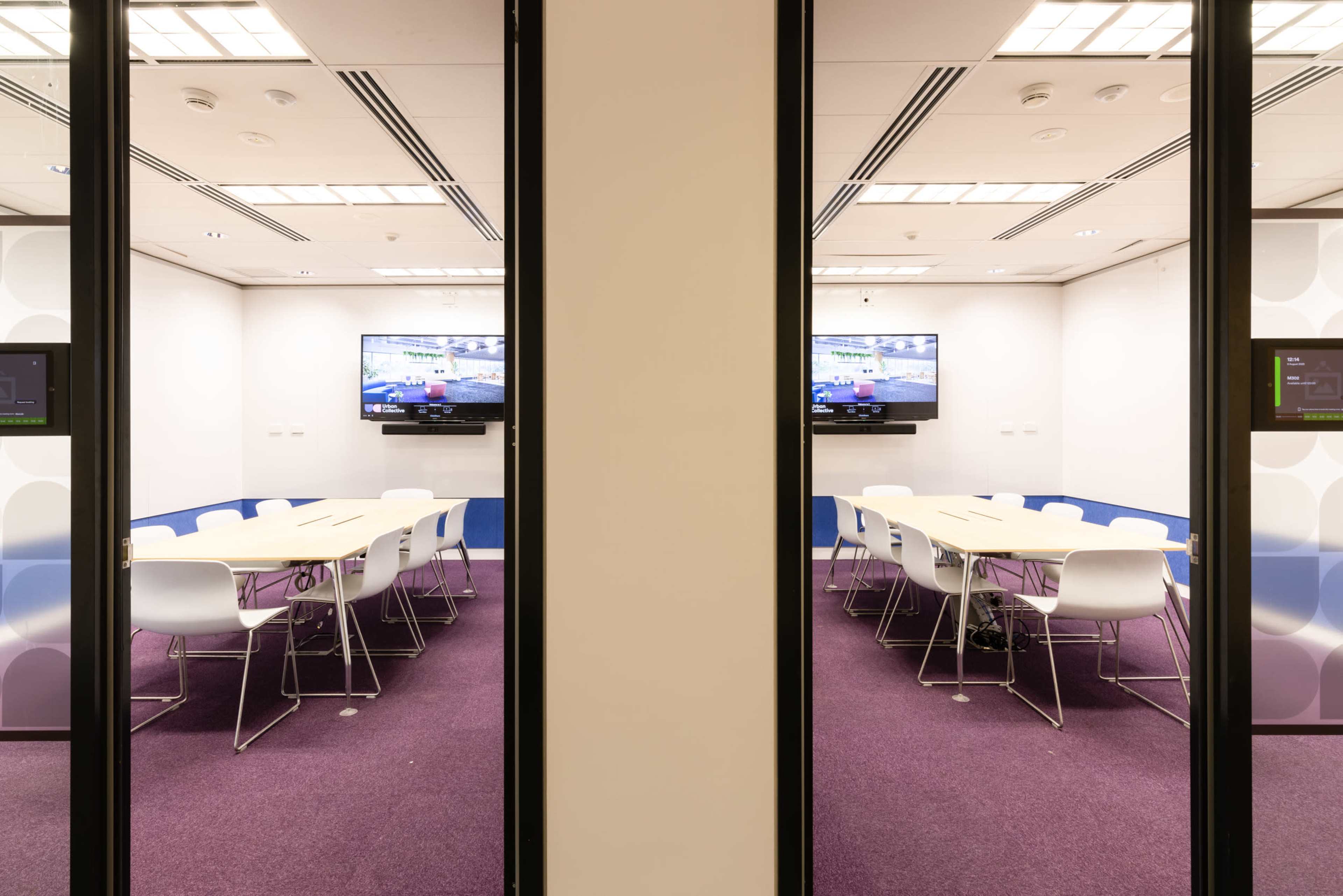 A modern conference room features a long table surrounded by white chairs, with two screens displaying content on opposite walls.
