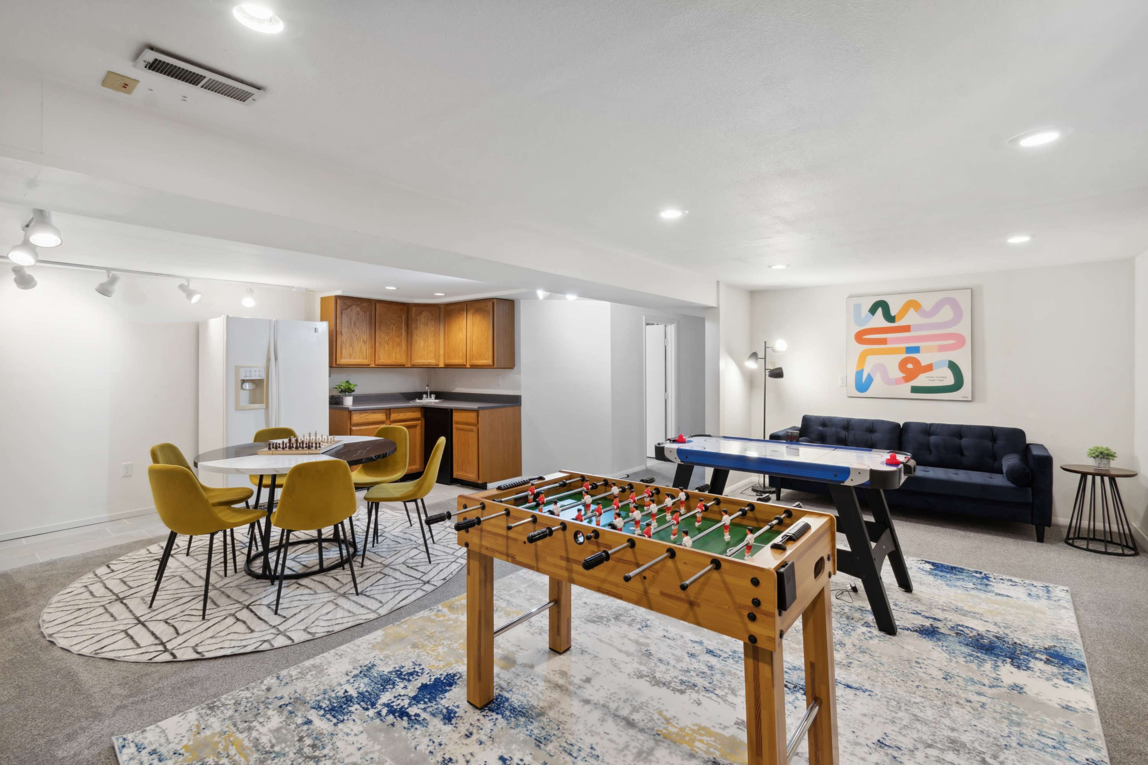 A finished basement features a foosball table, an air hockey table, a dining area with yellow chairs, and a sectional sofa.