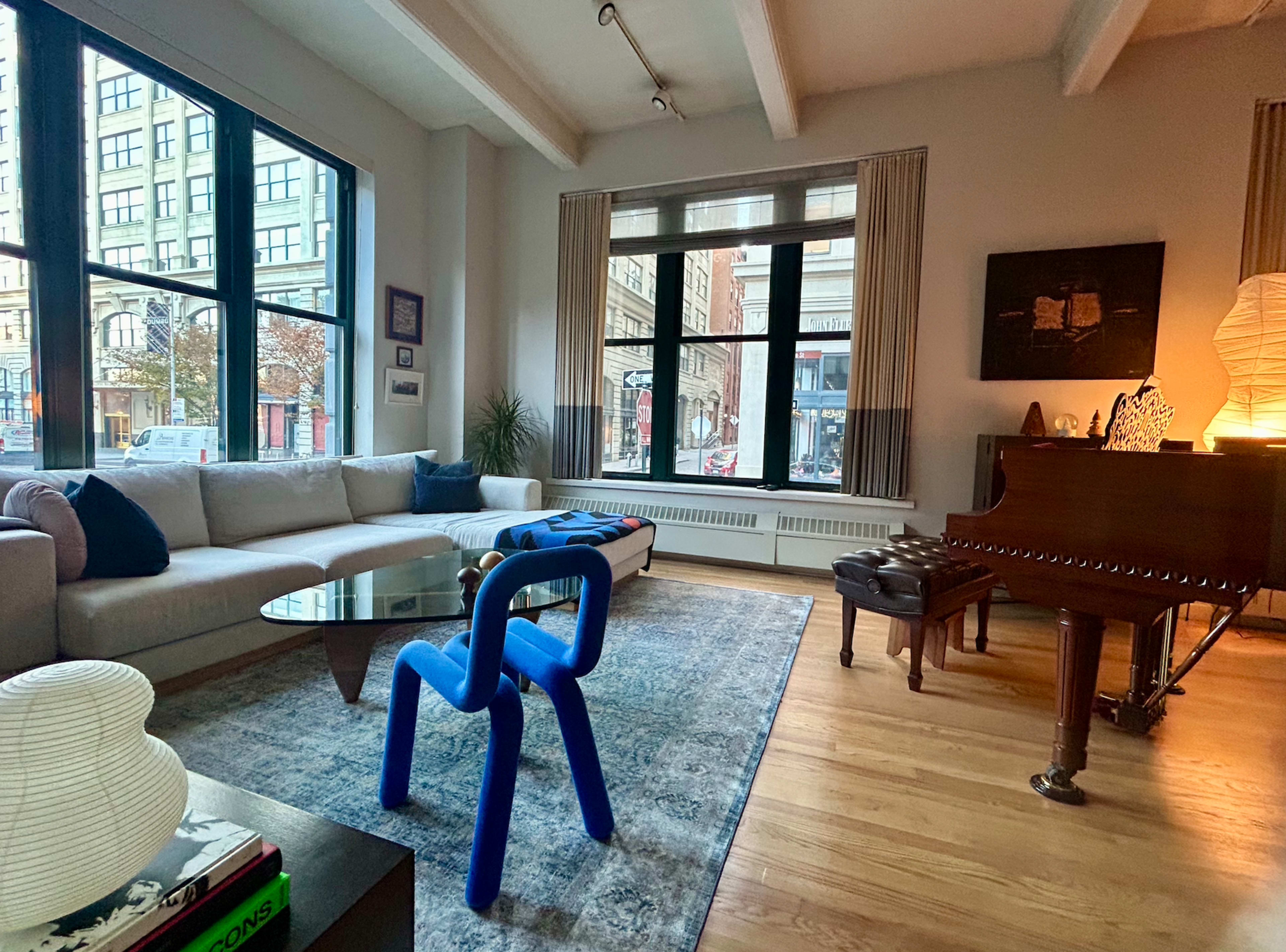A spacious living room features large windows, a gray sofa, a blue chair, a glass coffee table, and a grand piano.