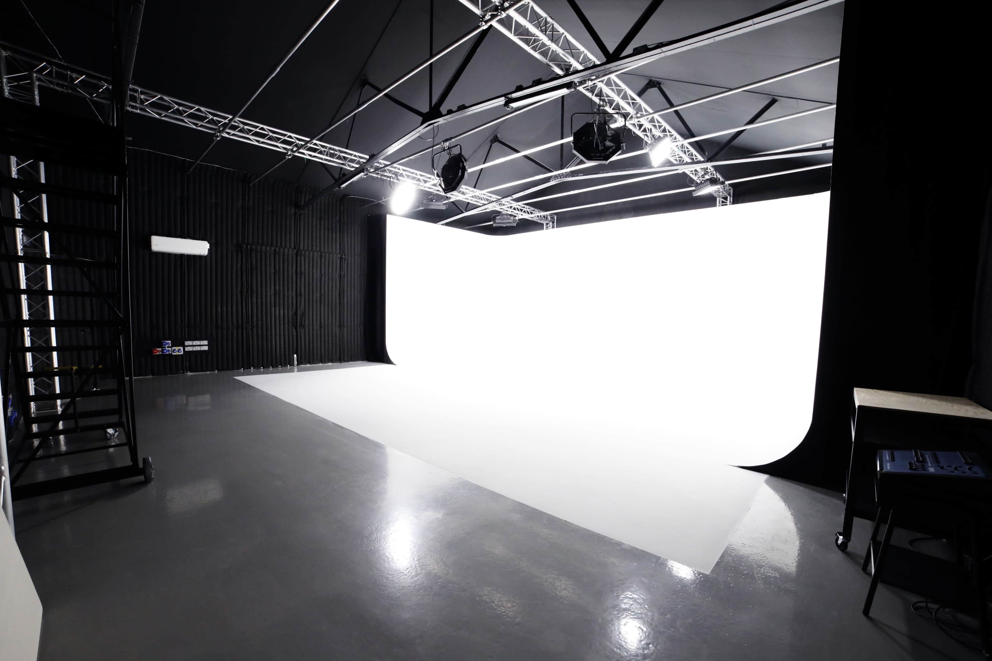 A photography studio features a large white backdrop, high ceilings, and overhead lighting.
