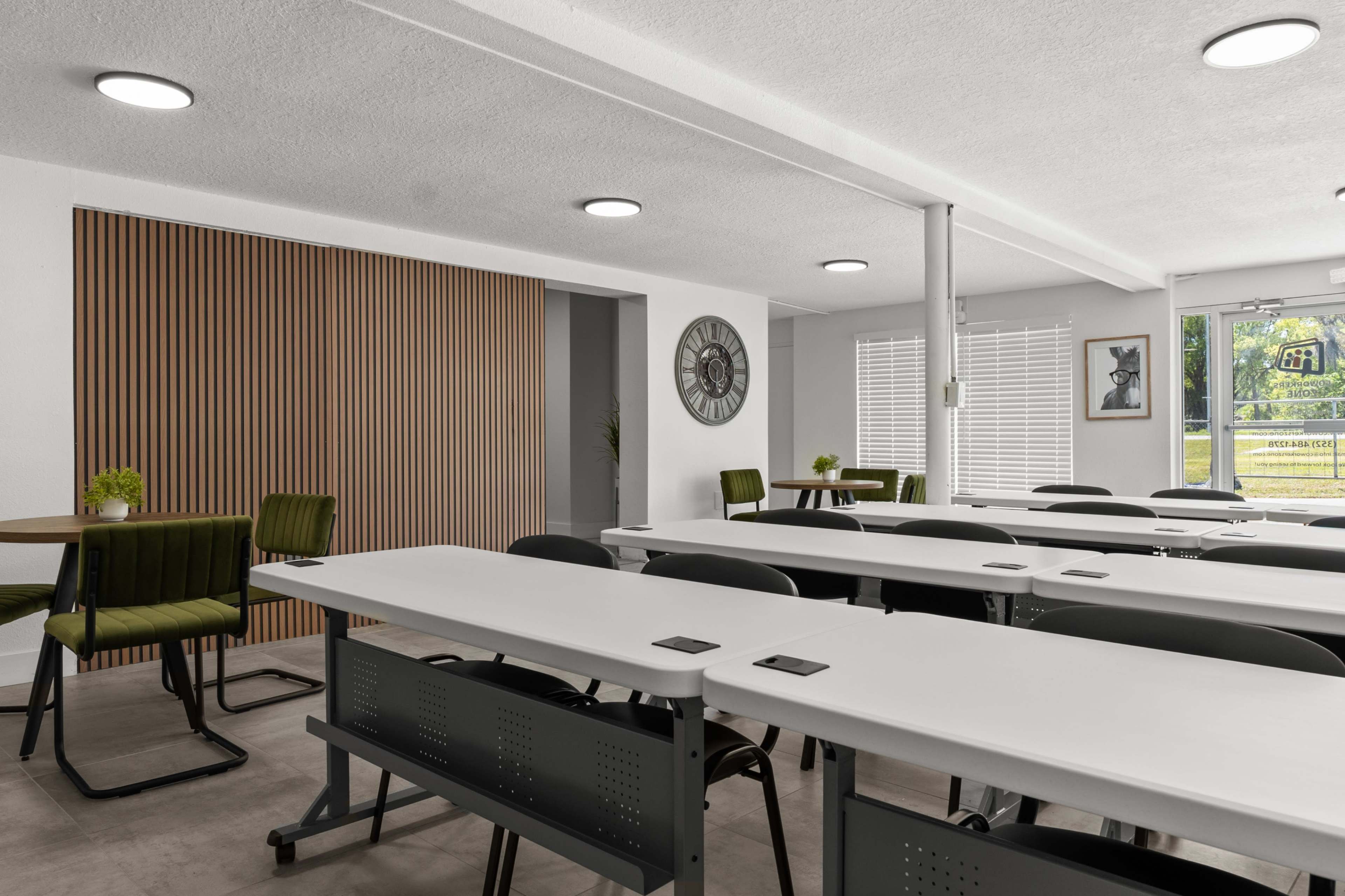 The image shows a modern, well-lit meeting room with rows of tables and chairs, a wall clock, and large windows.