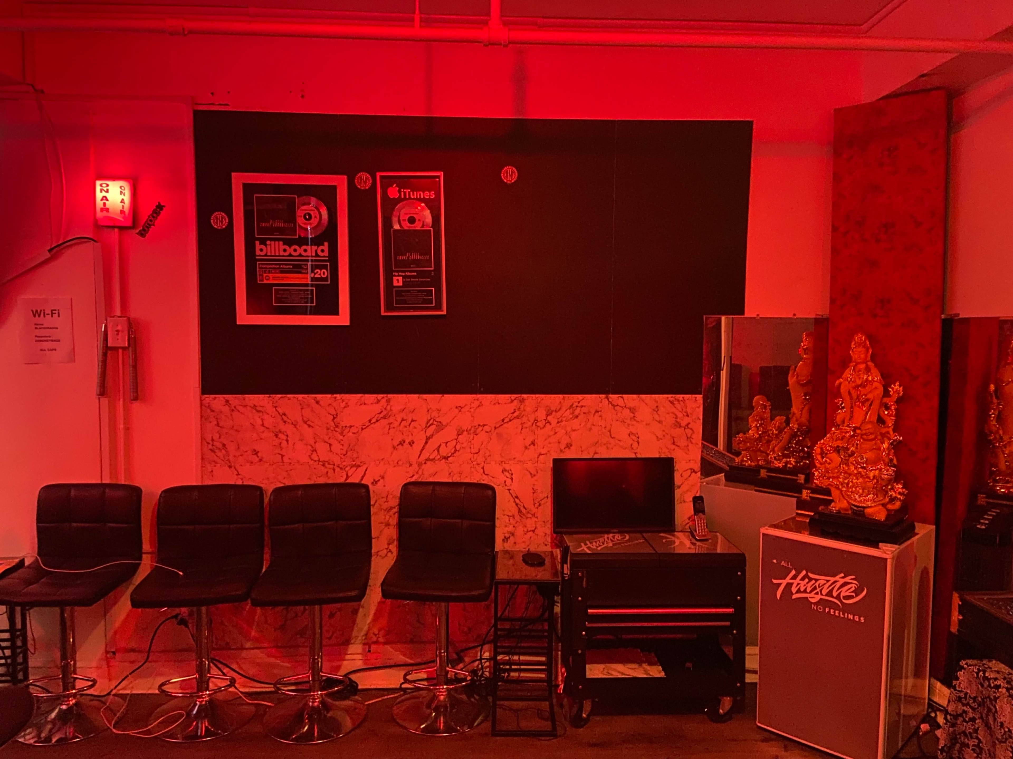 The image shows a dimly lit room featuring a black wall with music awards, several black bar stools, a small desk, a reflection of a statue in a mirror, and a promotional display in the corner.
