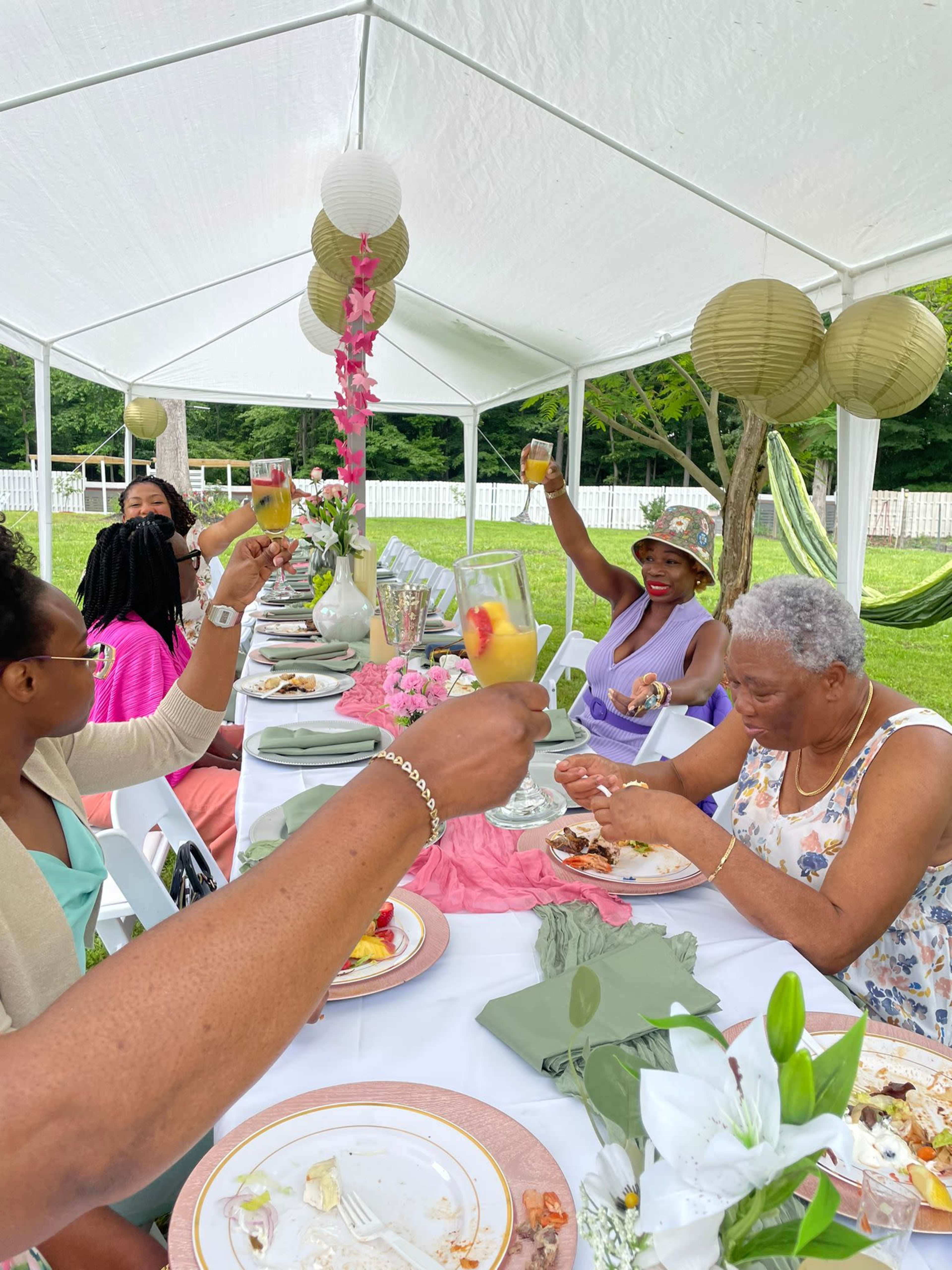 A group of people toasts with drinks at a festive outdoor gathering under a white tent, surrounded by tables set for a meal.