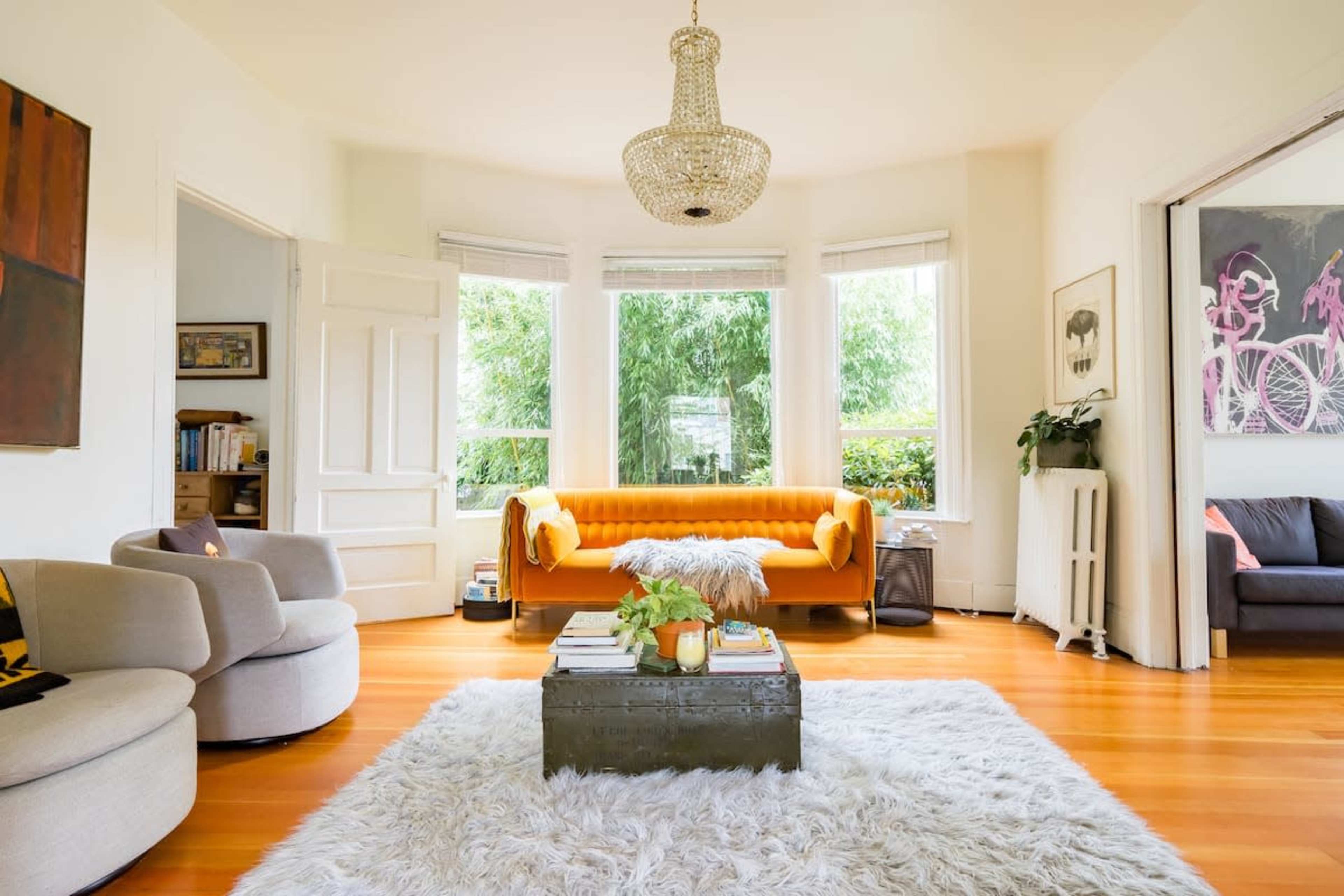 The scene depicts a bright living room featuring a yellow couch, a shaggy white rug, and large windows overlooking greenery.