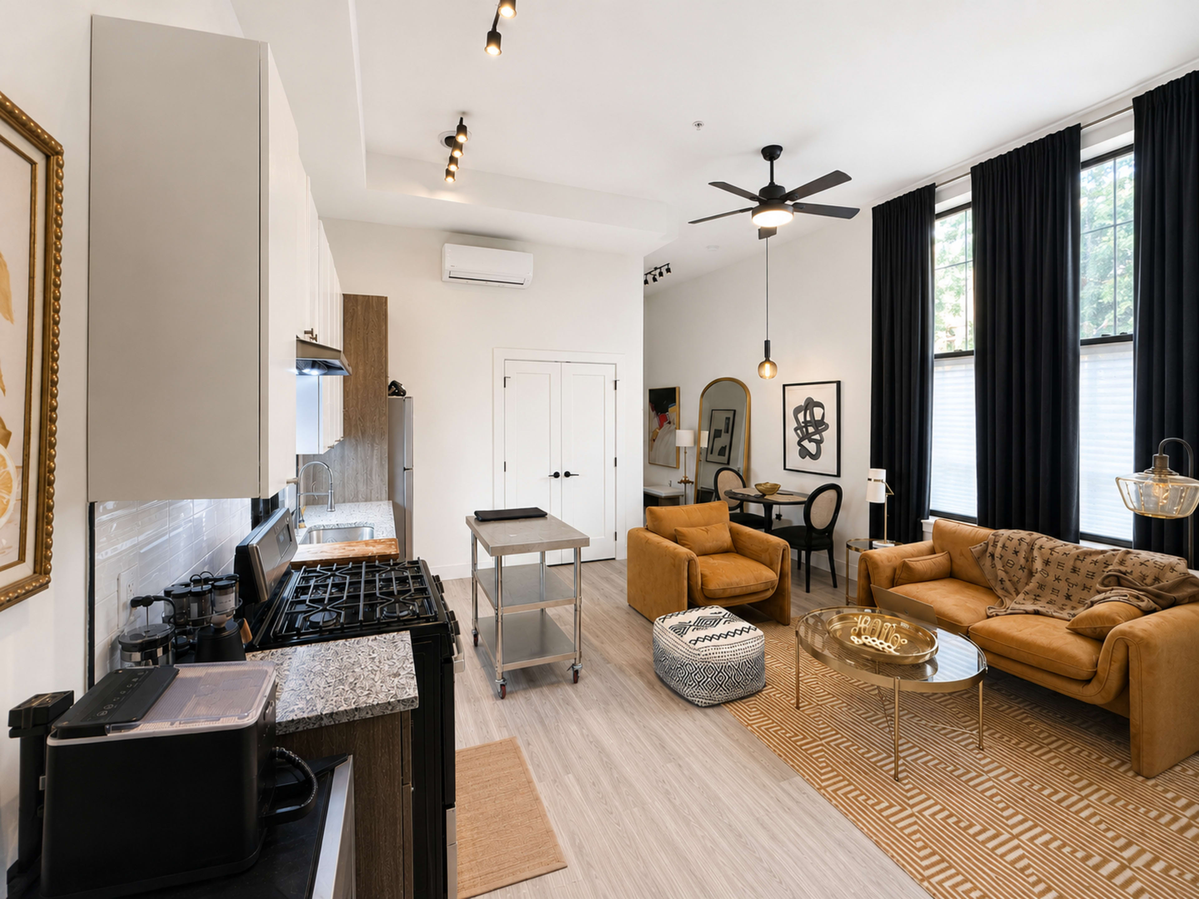 The image shows a modern, open-concept living space featuring a kitchen with black appliances, a living area with brown sofas, a round glass coffee table, and dining space with a circular table and chairs.