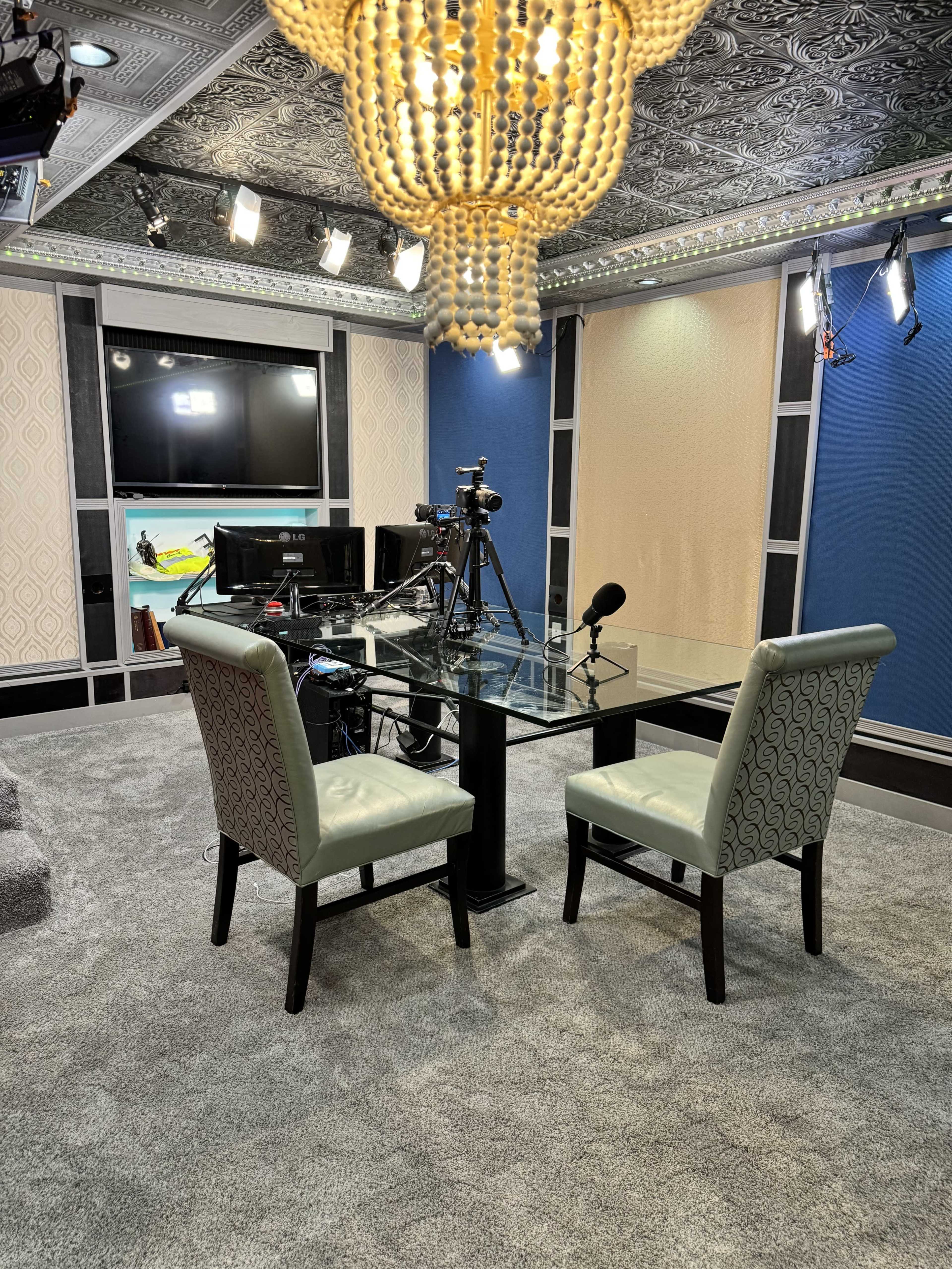 The image shows a modern recording studio featuring a glass table, two upholstered chairs, and professional camera equipment on a tripod.