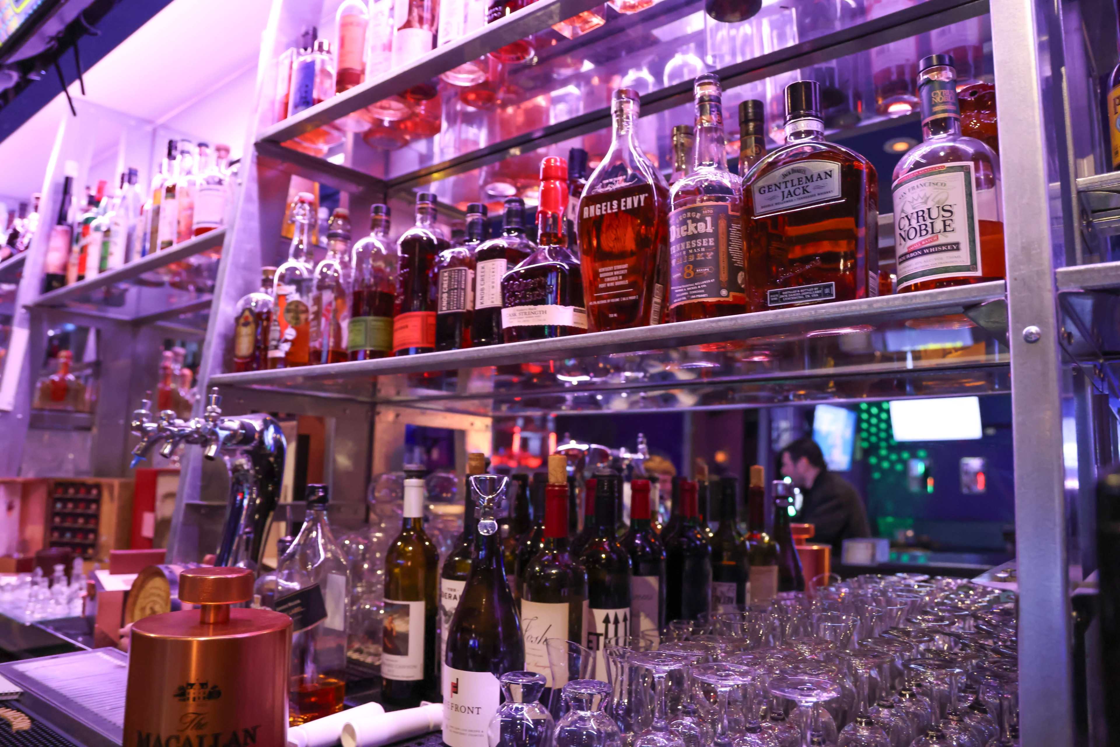 The image shows a well-stocked bar shelf displaying a variety of liquor bottles, including whiskey and wine, with glasses arranged neatly below.