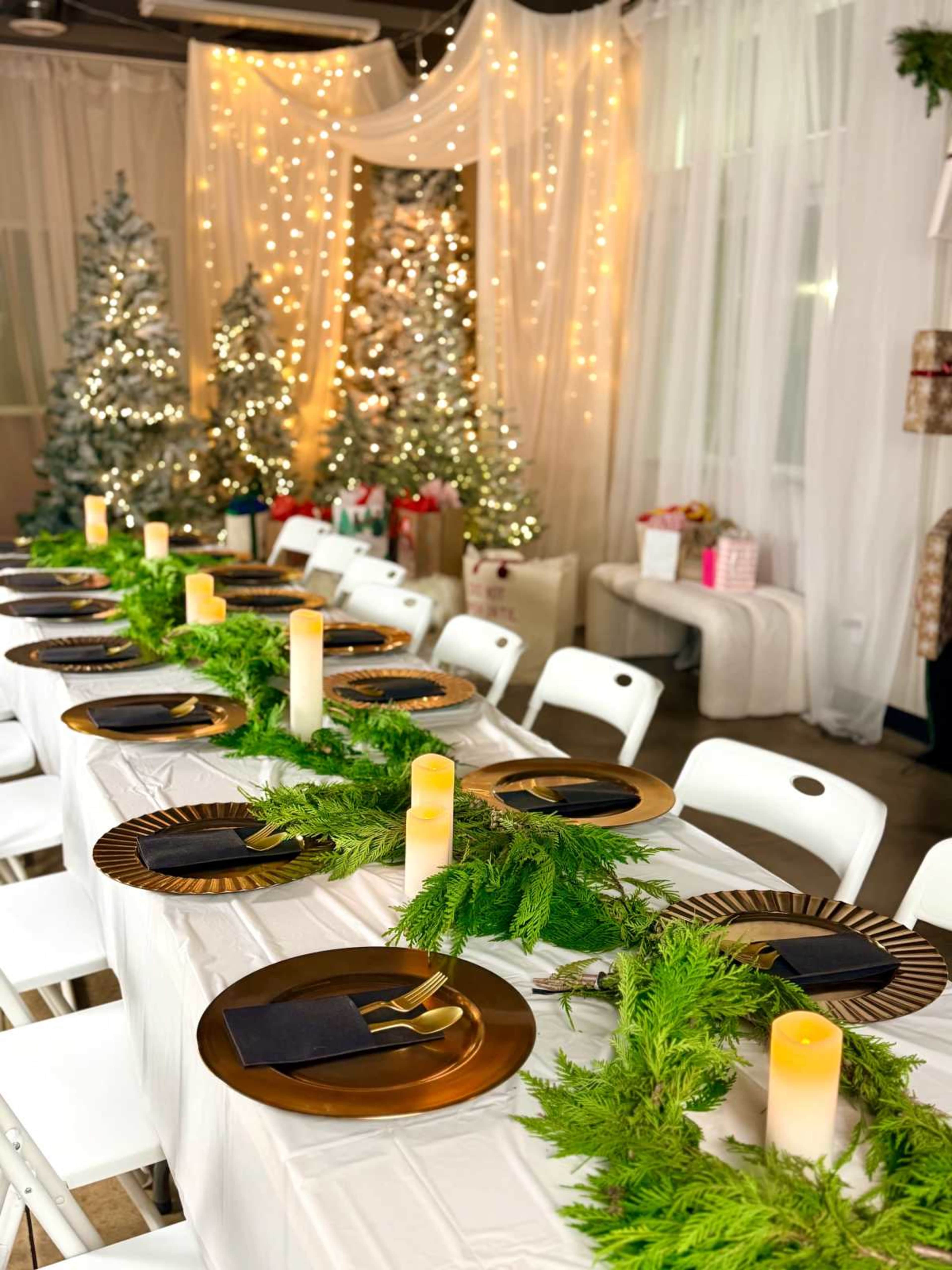 A long dining table is set with gold plates and dark napkins, adorned with a green garland and surrounded by decorated Christmas trees in a festive room.